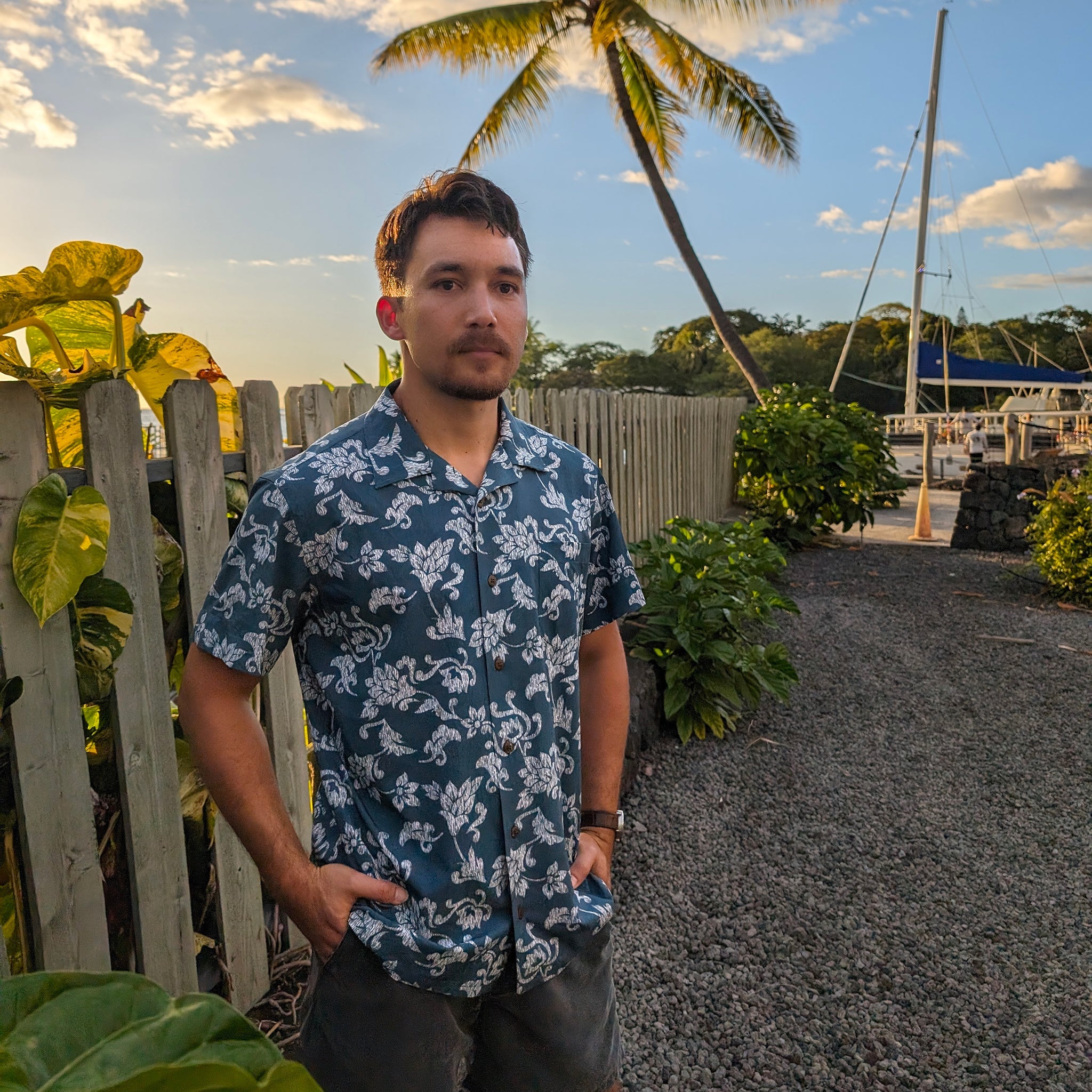 Man wearing a blue floral shirt standing in a tropical setting with palm trees and boats.