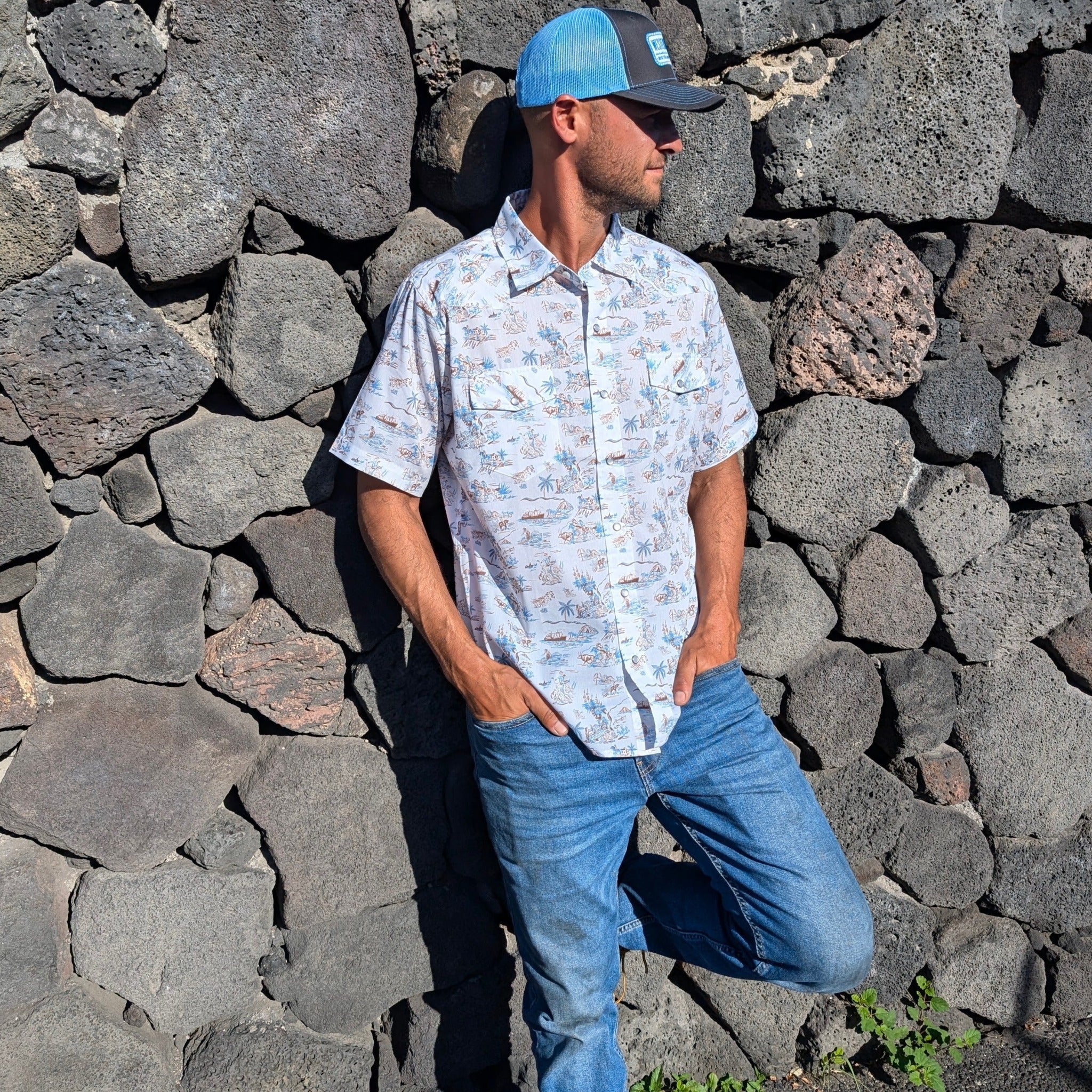 Man wearing a light-colored patterned shirt and blue jeans standing against a stone wall.