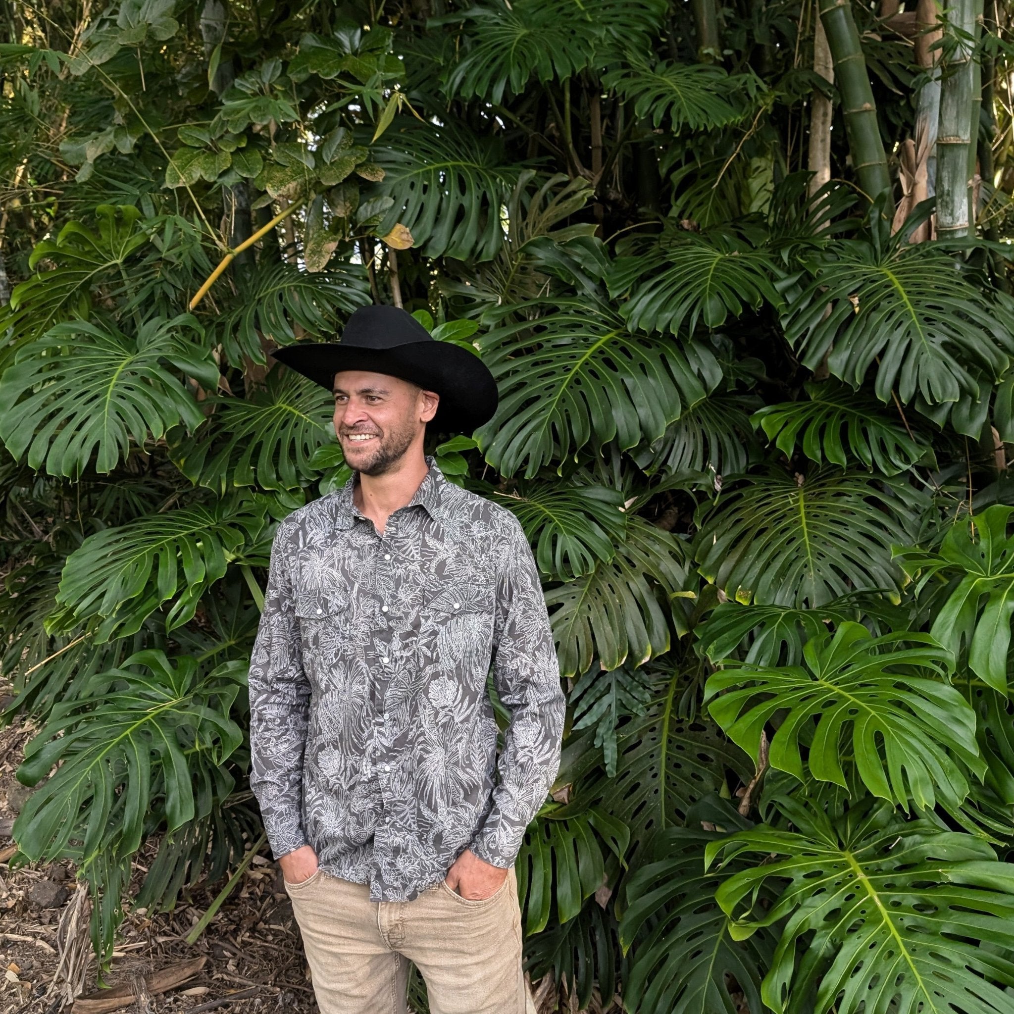 Man wearing a black cowboy hat and patterned shirt standing in front of lush green foliage