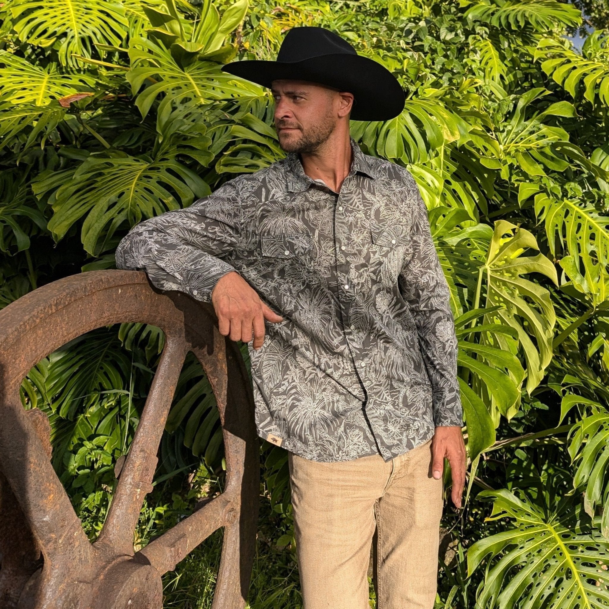 Man wearing a patterned shirt and cowboy hat standing in front of green foliage.