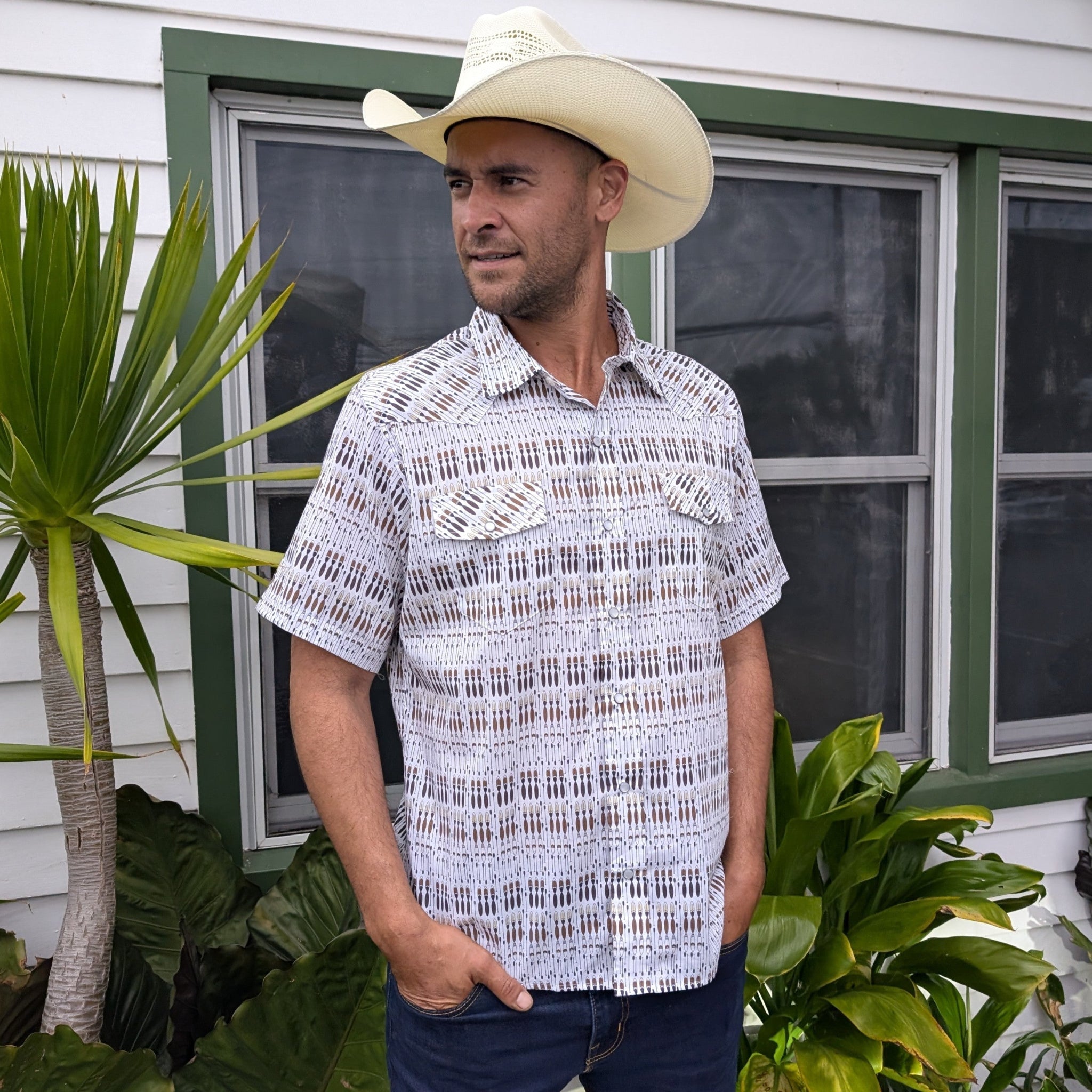 Man wearing a cowboy hat and patterned shirt standing in front of a house with plants.