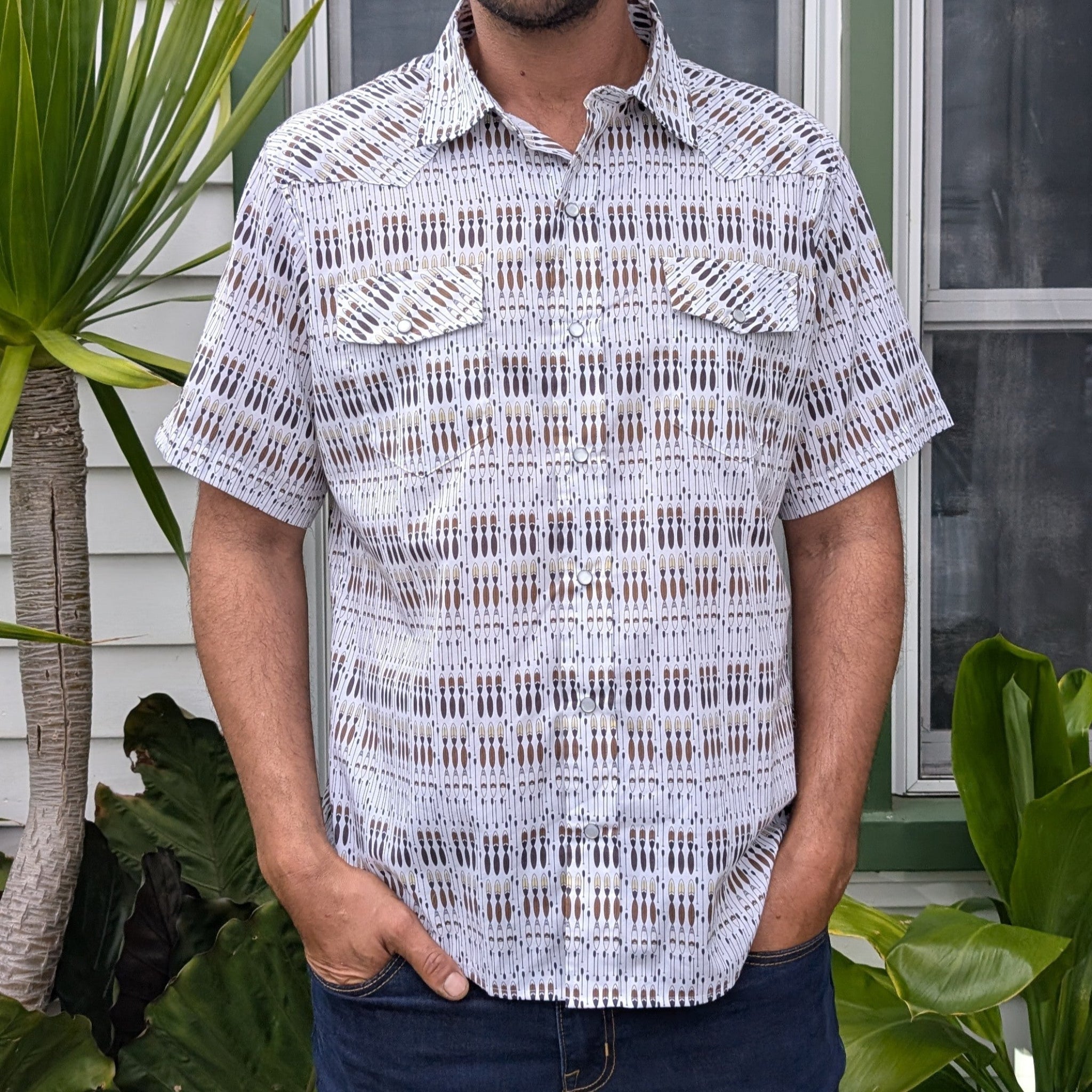 Person wearing a patterned short-sleeve shirt standing outdoors with plants in the background