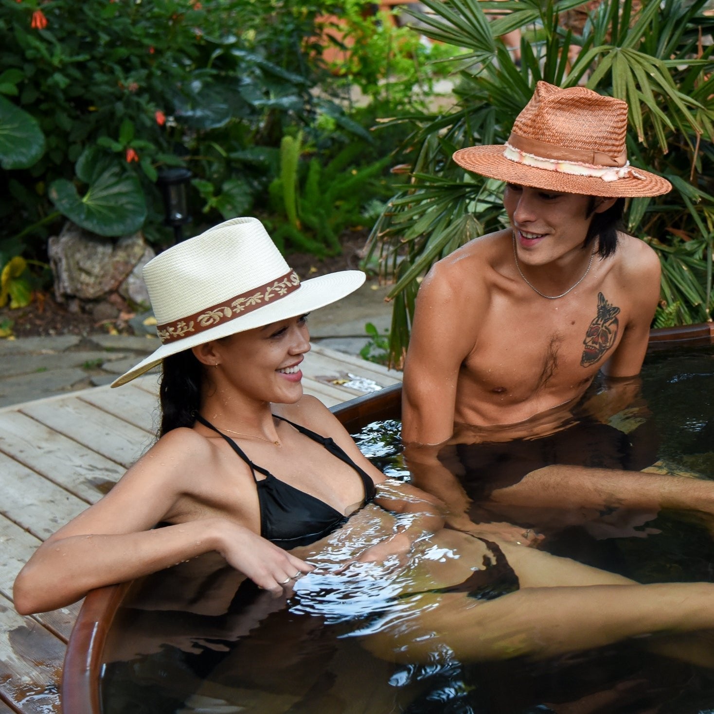 Two people sitting in a hot tub with hats on, surrounded by greenery.