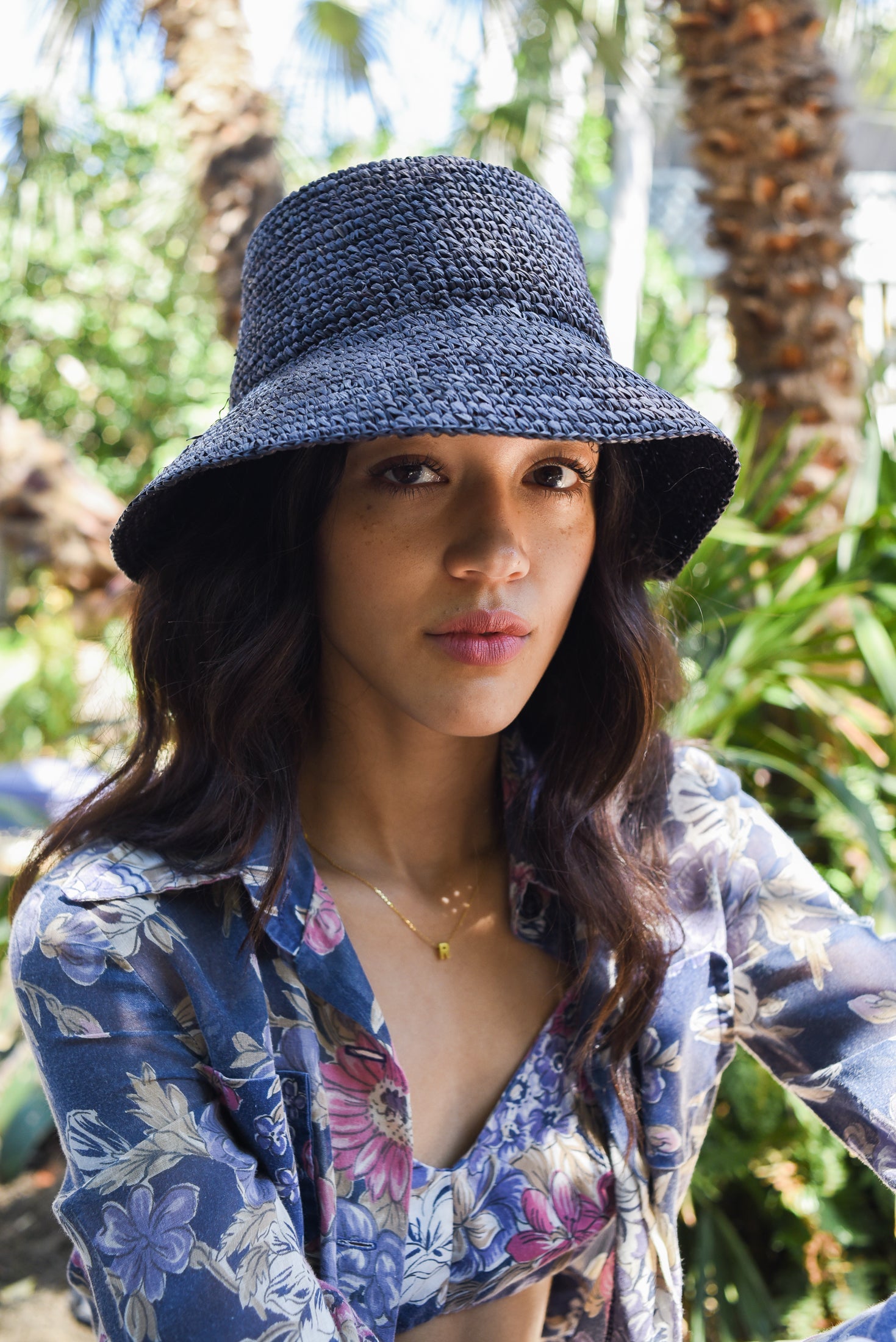 Woman wearing a blue sun hat and floral shirt outdoors with palm trees in the background
