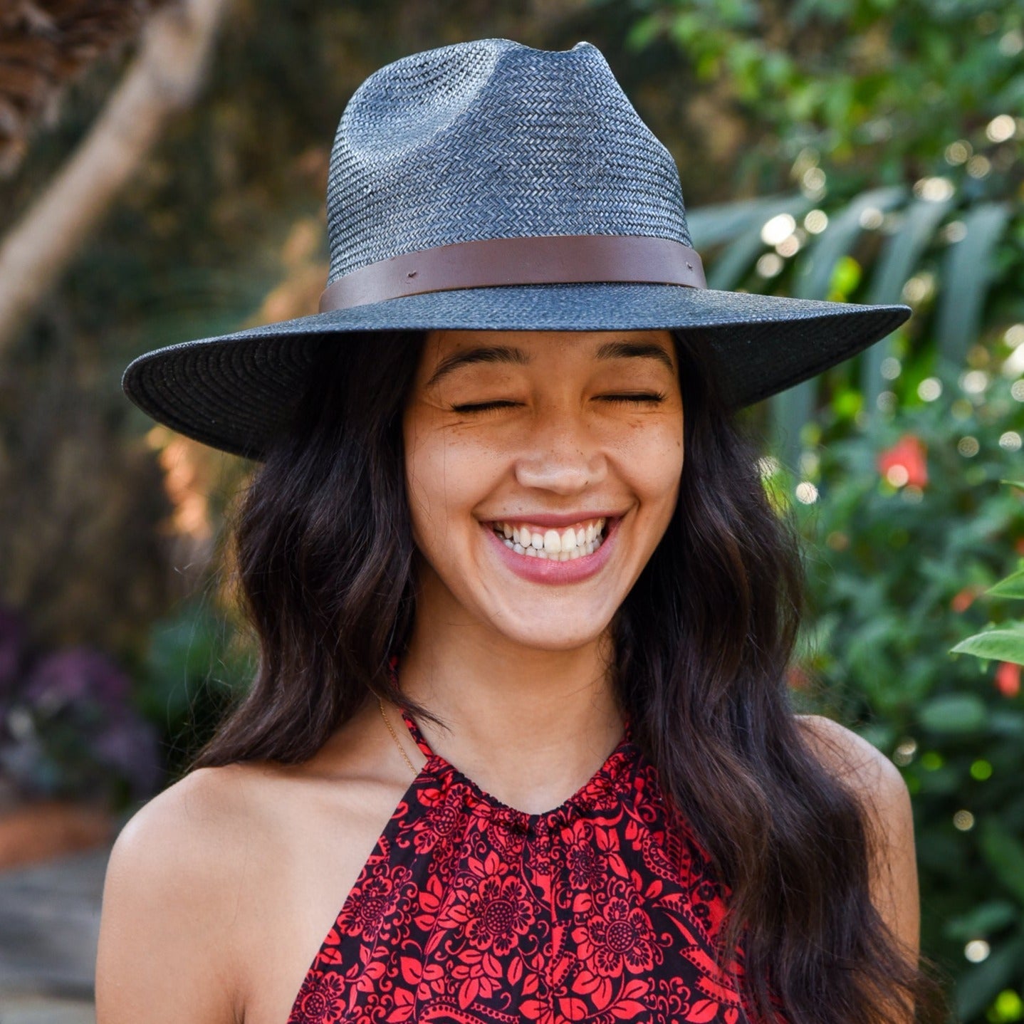 Woman wearing a blue fedora with a brown band, smiling outdoors with greenery in the background.