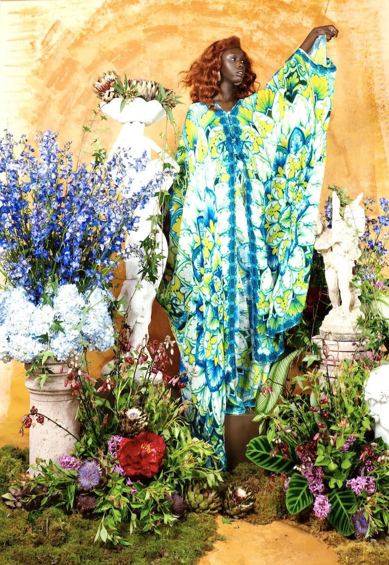 Woman in a colorful dress standing among flowers and statues against a yellow wall.