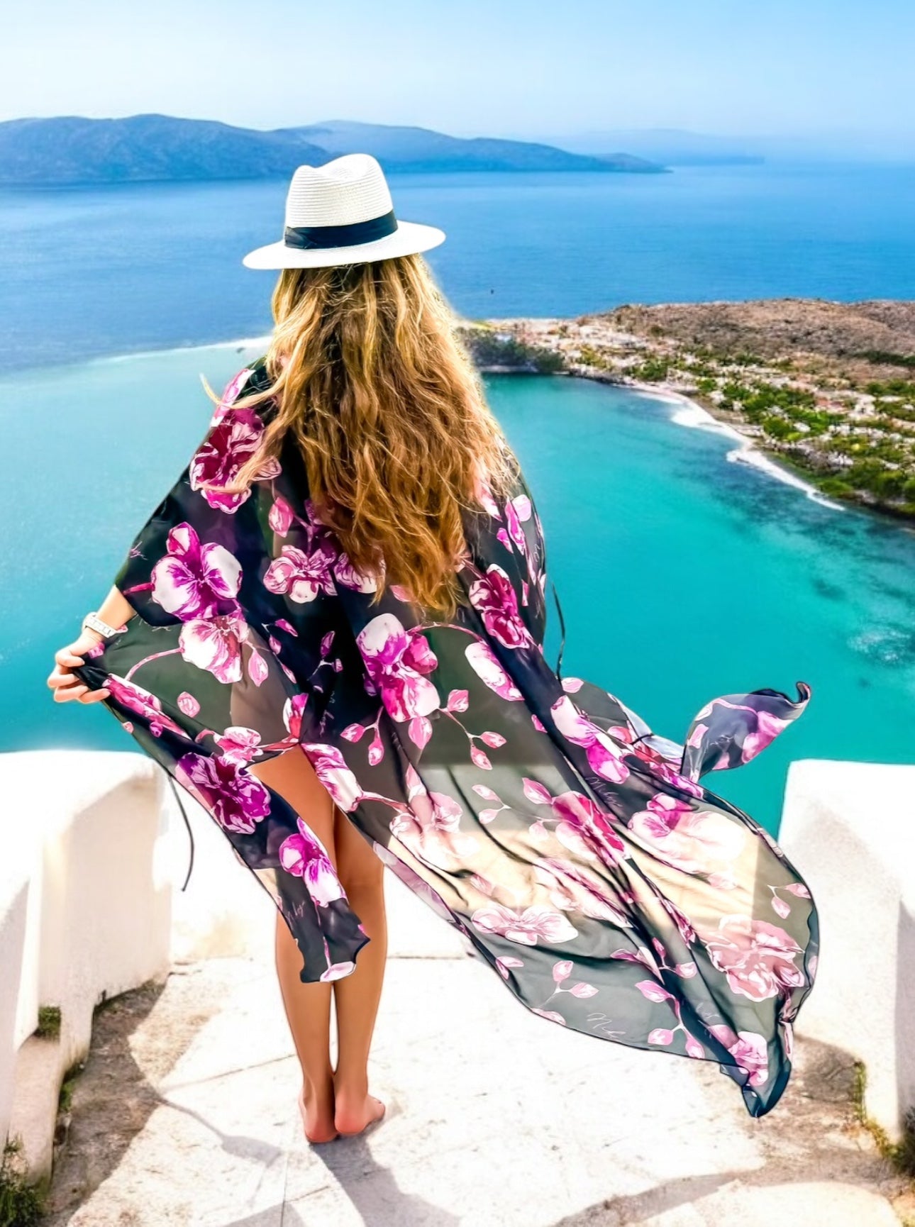 Woman in a floral kimono and hat overlooking a scenic ocean view.
