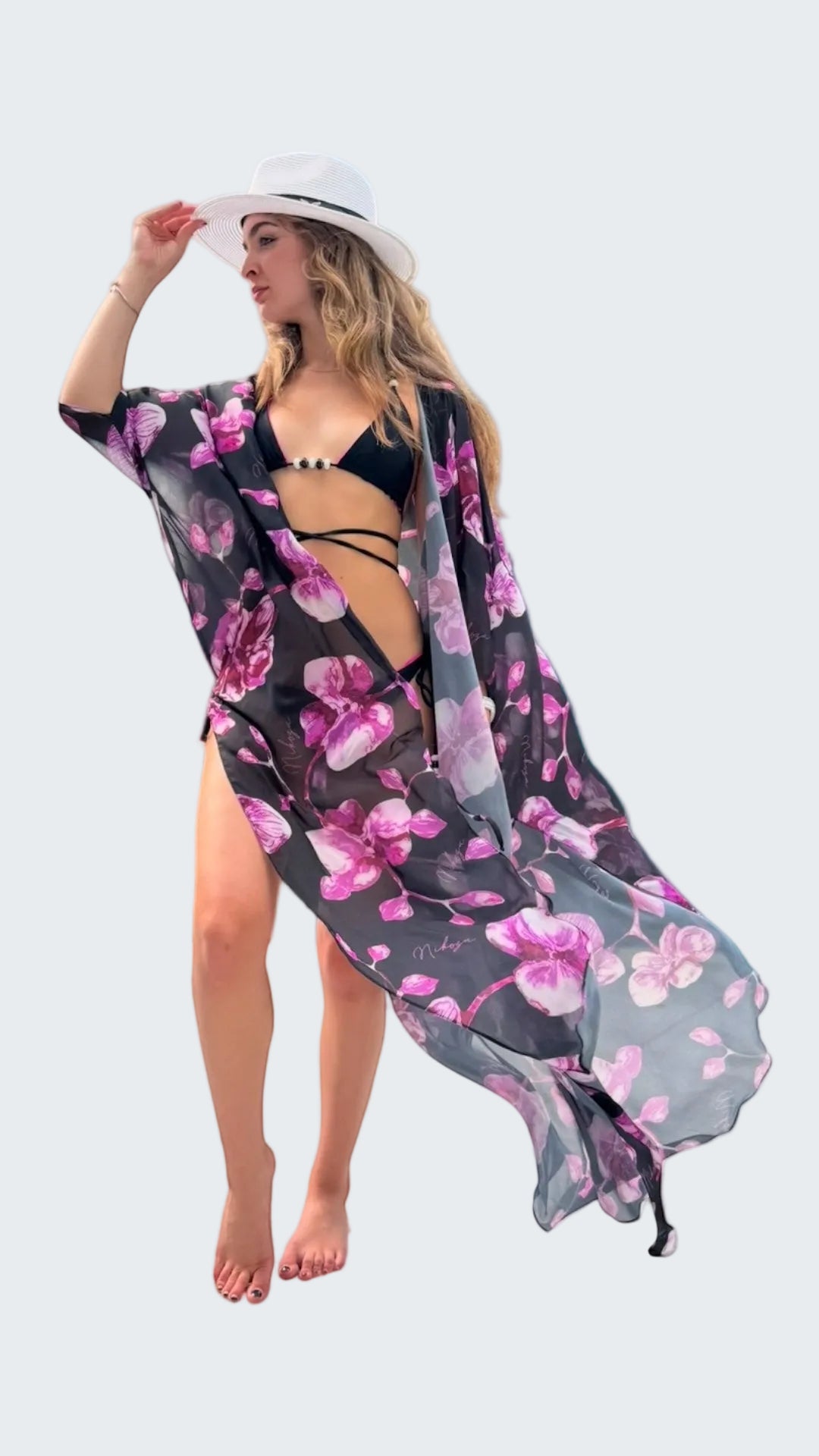 Woman in a floral kimono and black bikini standing on a rocky shore with ocean and sky in the background.