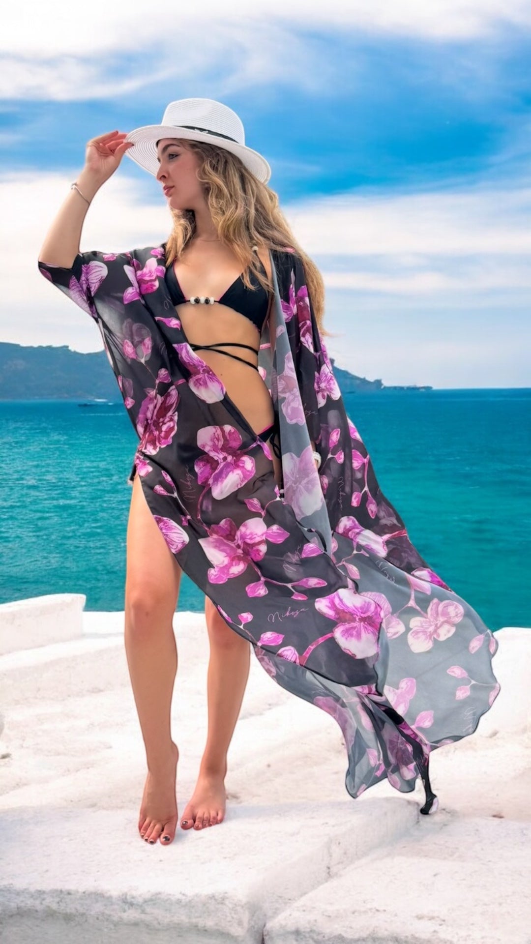 Woman in a floral kimono and black bikini standing on a rocky shore with ocean and sky in the background.
