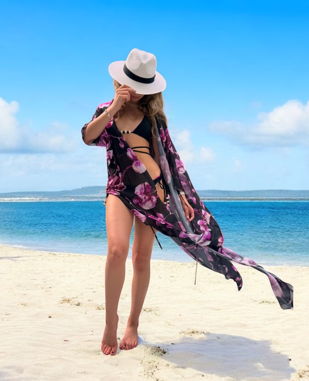 Woman in a floral cover-up and hat on a beach with clear blue sky and ocean.