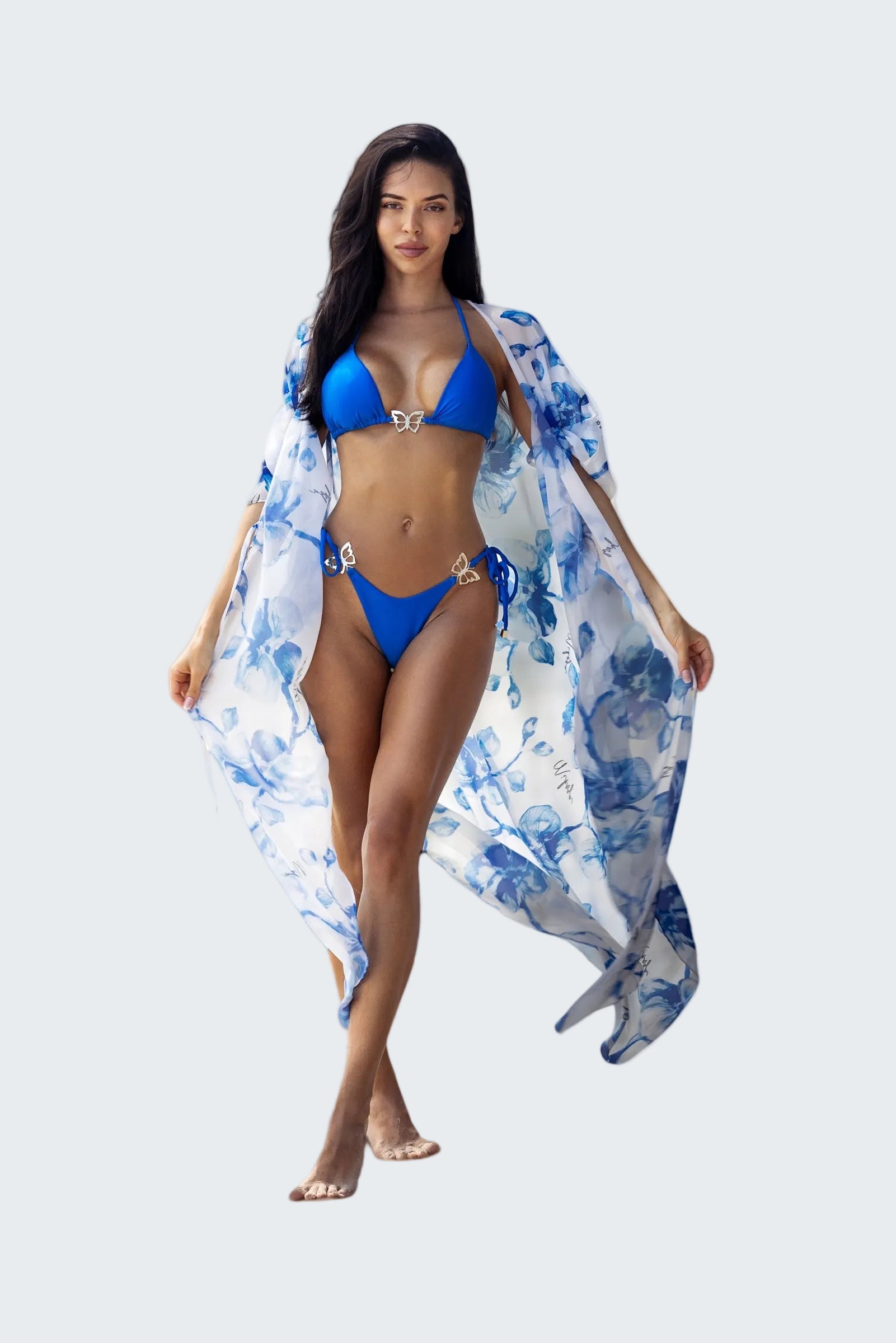 Woman in a blue bikini with a floral cover-up standing on a beach with ocean view.