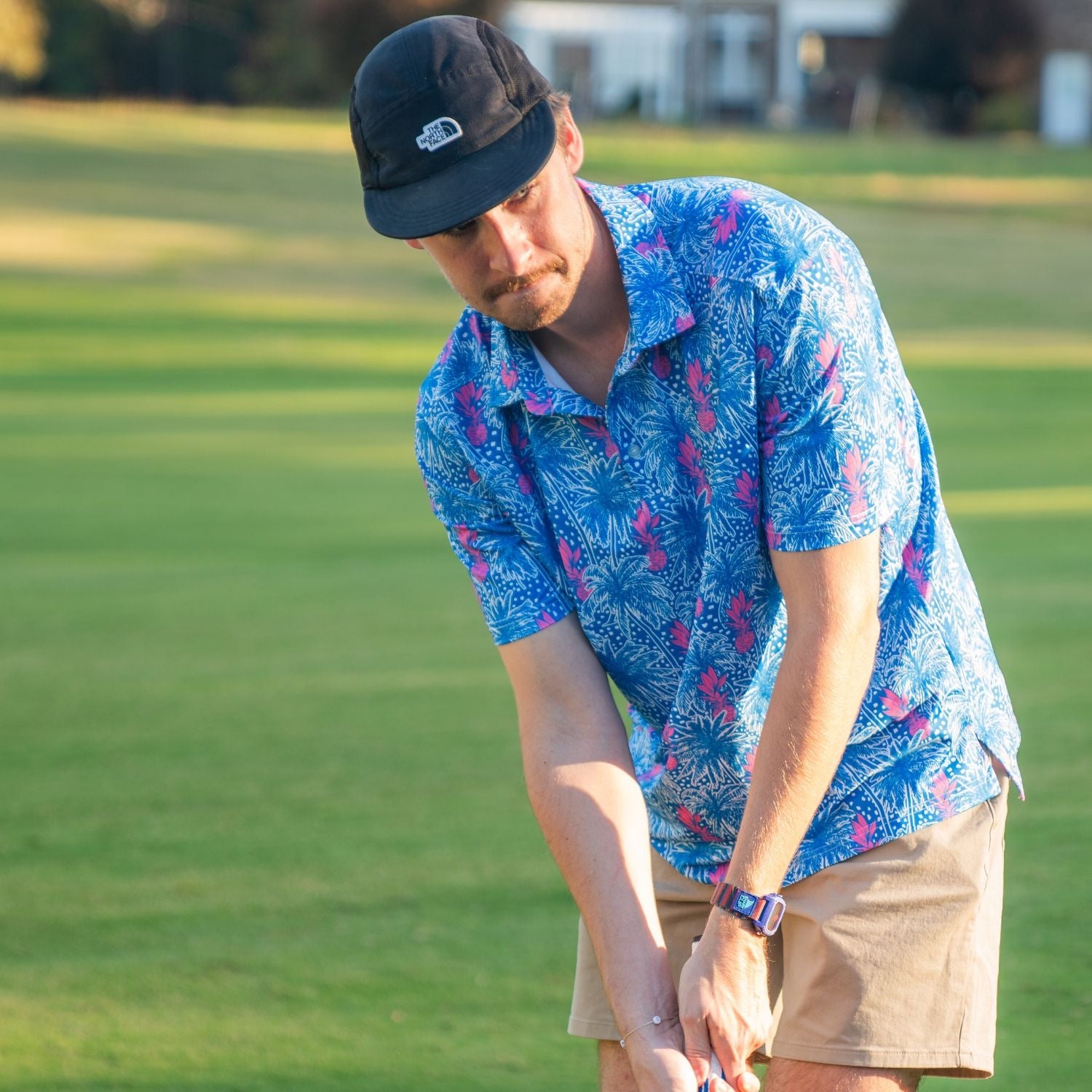 Man in a blue floral shirt and black cap playing golf on a green field.
