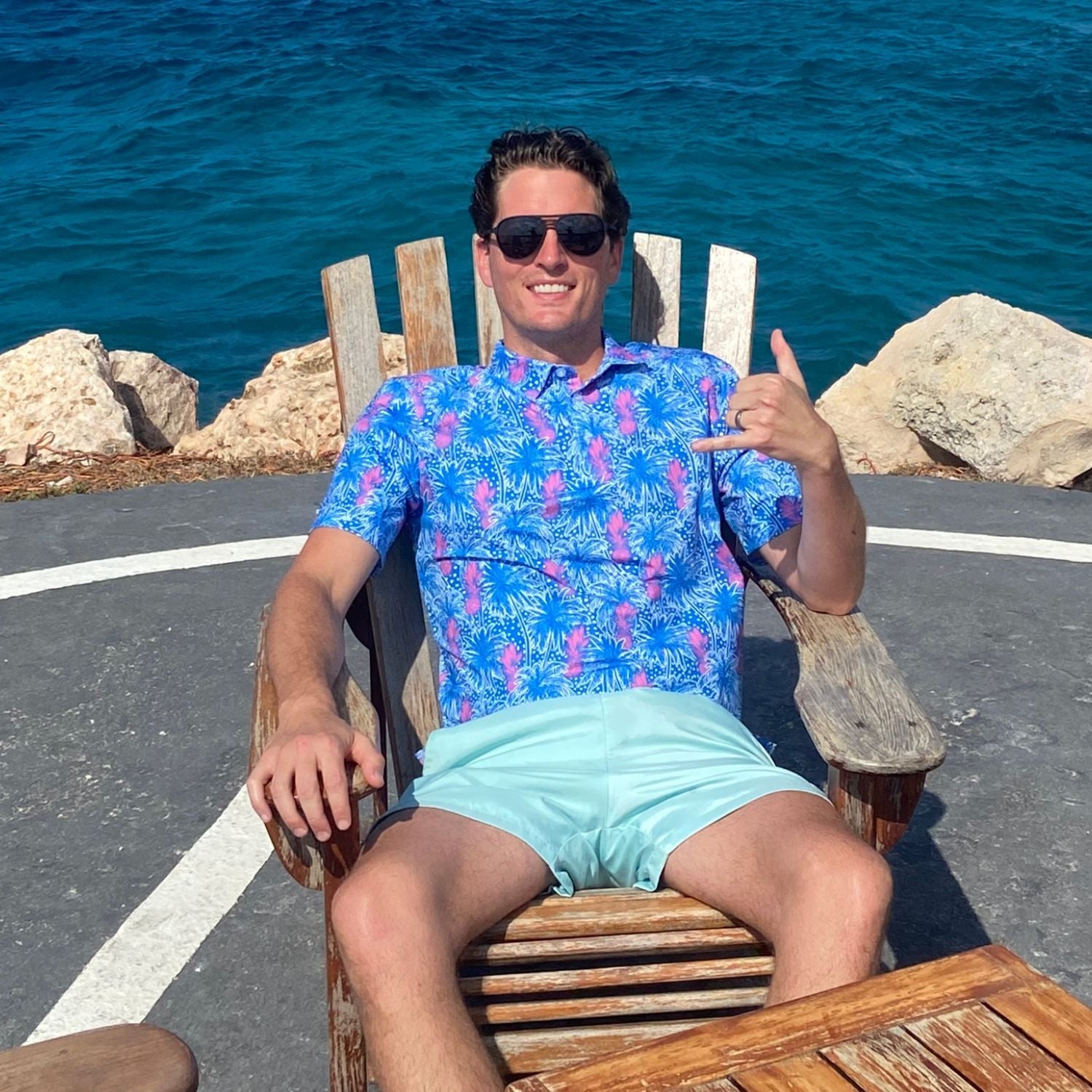 Man sitting on a wooden chair by the sea, wearing sunglasses and a blue patterned shirt.