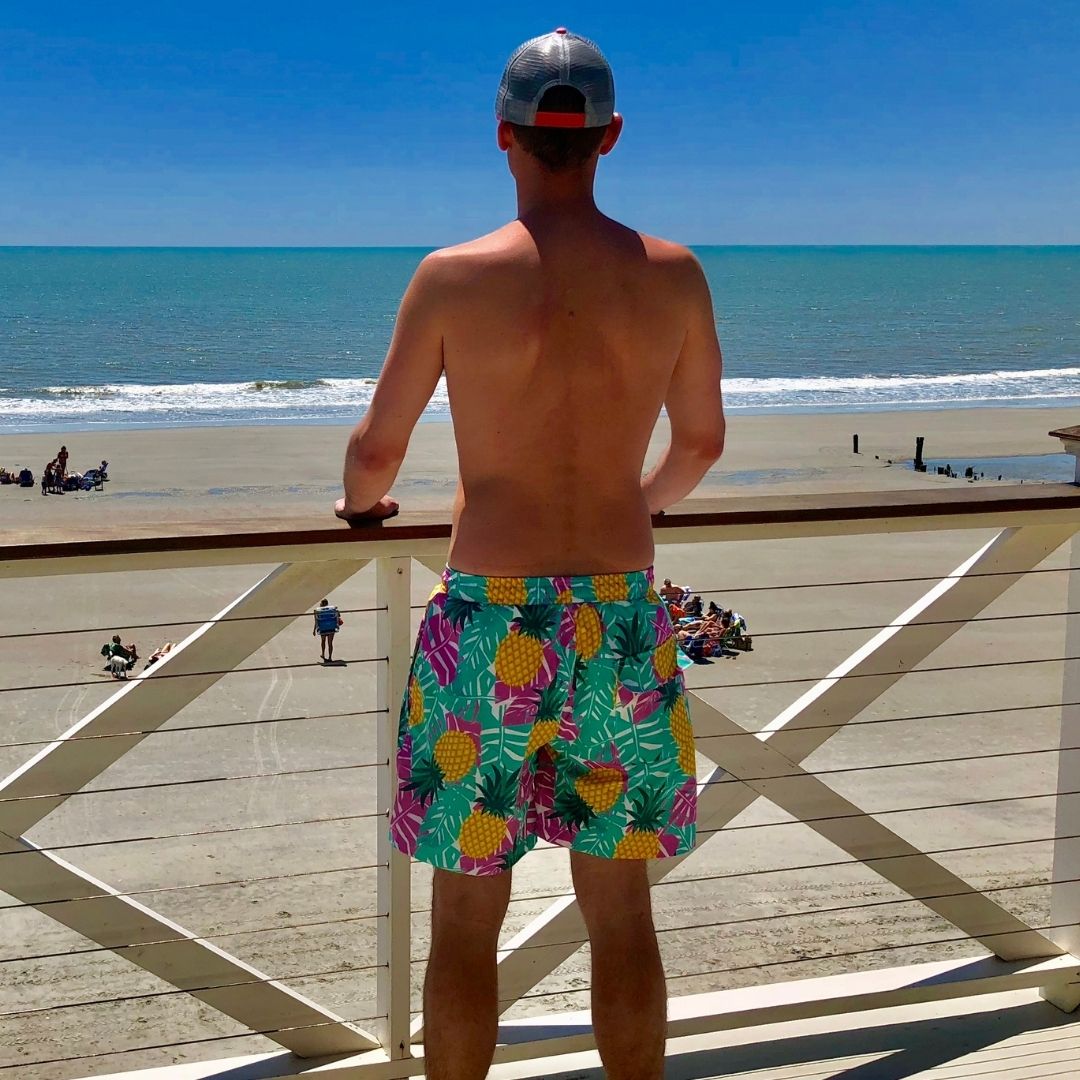 Man wearing colorful pineapple-patterned shorts standing on a wooden deck overlooking a beach.