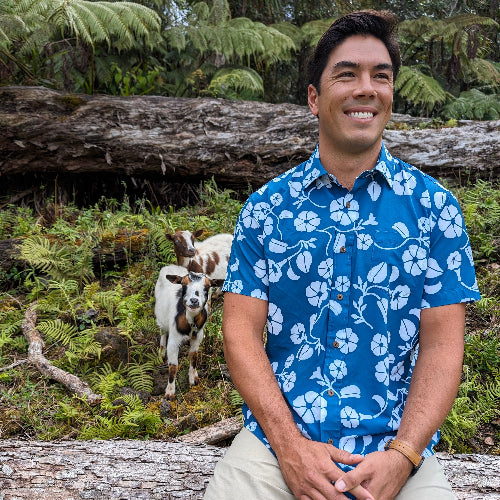 Man in a blue floral shirt sitting on a log with goats in a forest setting