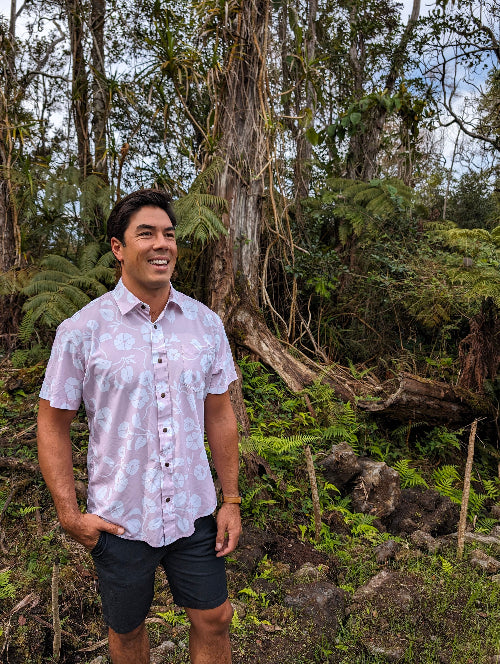 Man standing in a forested area wearing a patterned shirt and shorts.