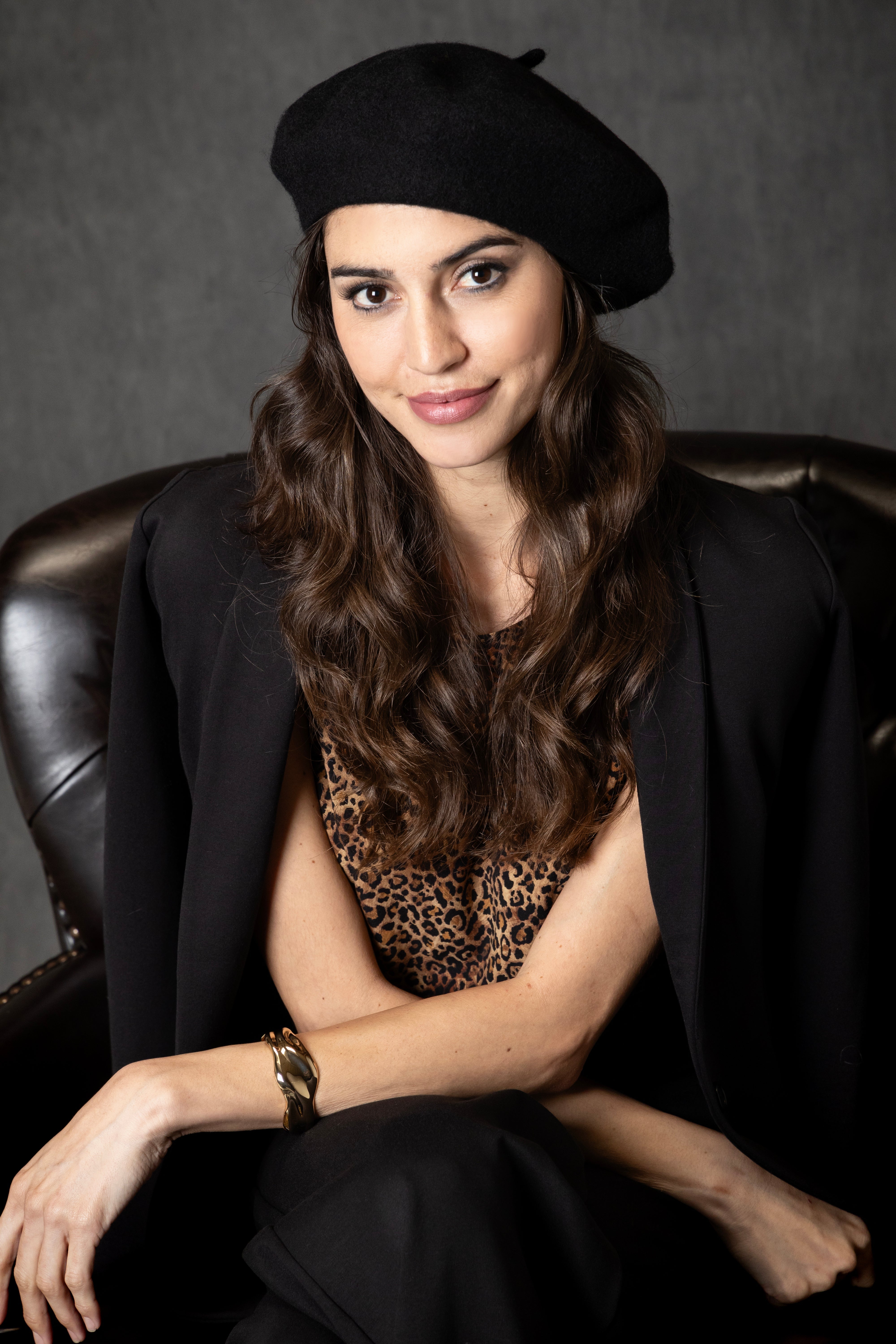 Woman wearing a black beret and black coat with a leopard print top, sitting on a leather chair.