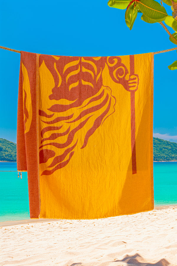 Towel with a lion design hanging on a beach with clear blue water and sky.
