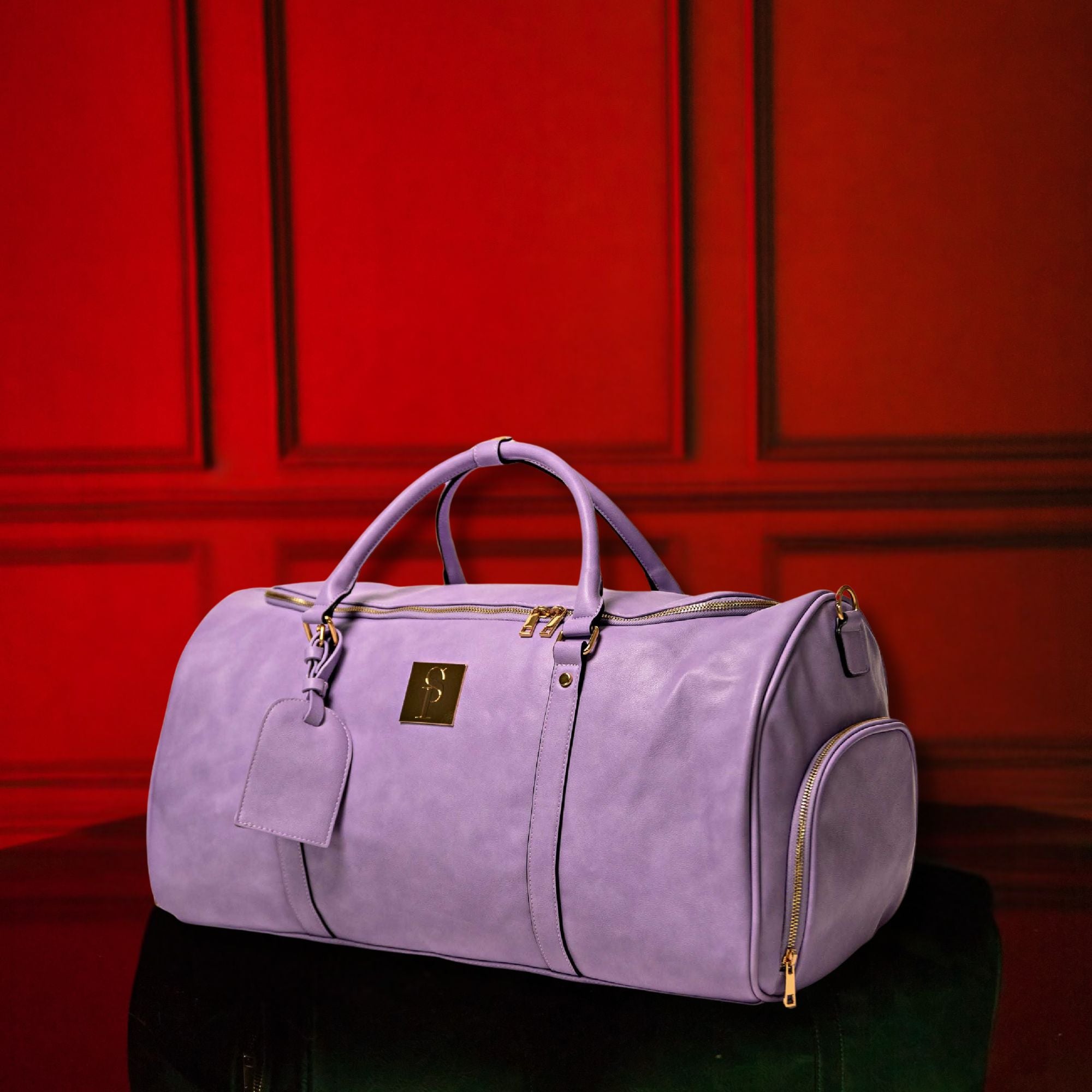 Purple duffel bag with gold accents on a reflective surface against a red paneled wall.