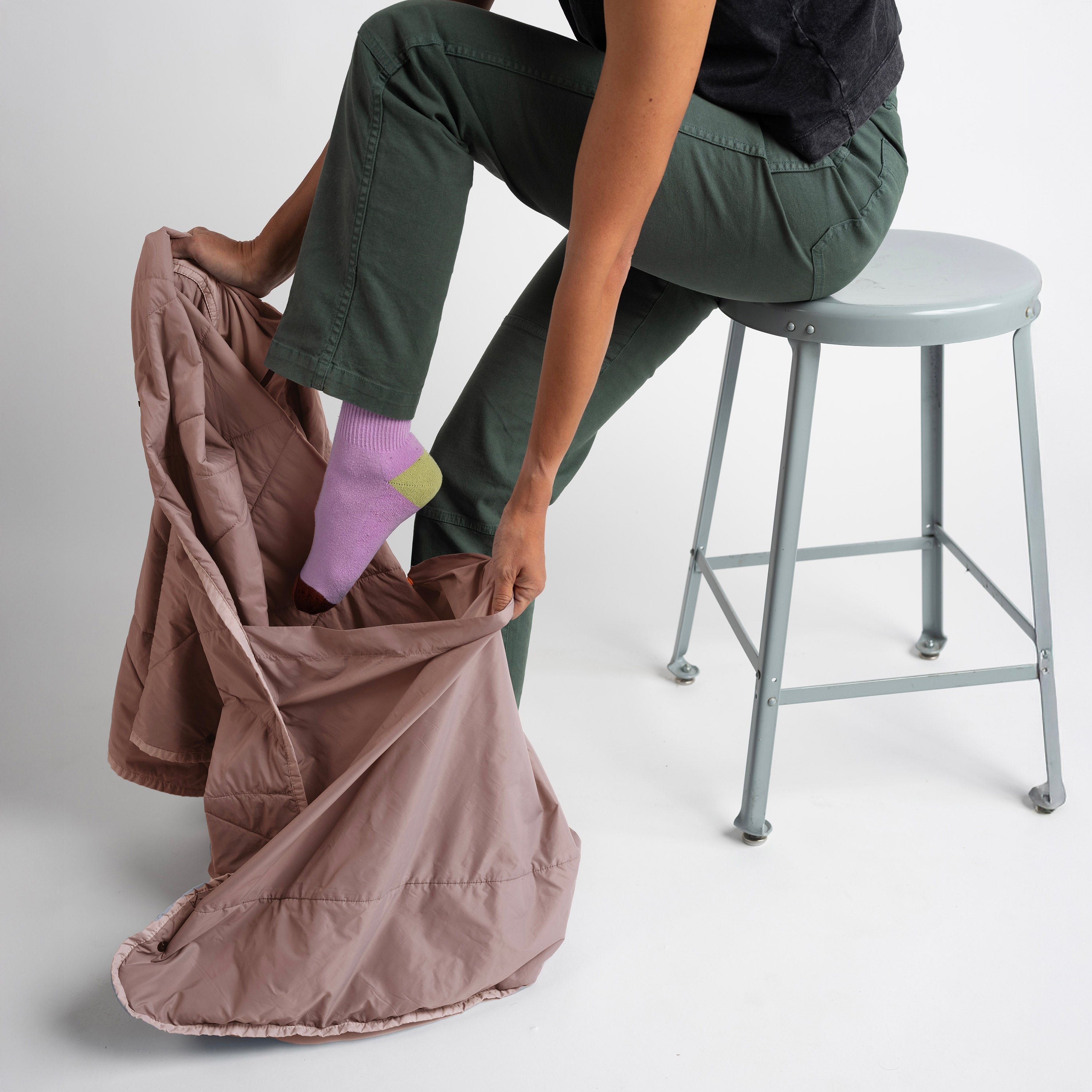 Person sitting on a stool with one leg inside a brown bag, wearing green pants and pink socks.