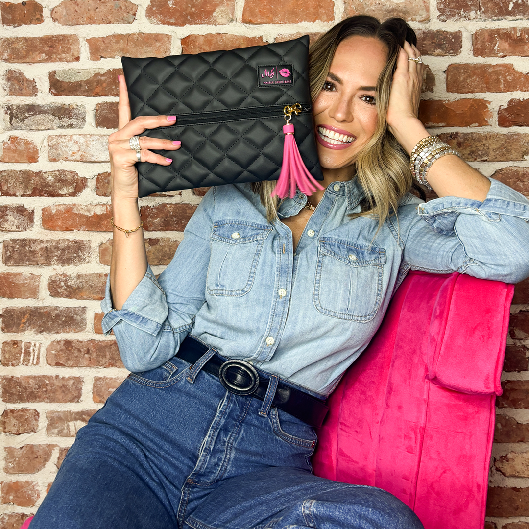Woman holding a black quilted clutch with a pink tassel against a brick wall.