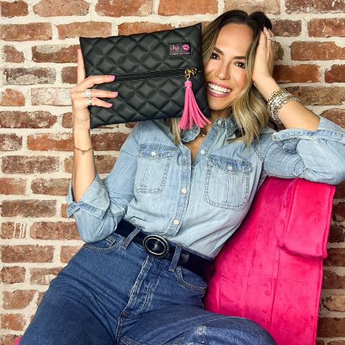 Woman holding a black quilted clutch with a pink tassel against a brick wall.