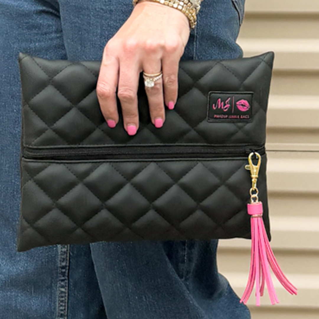 Black quilted makeup bag with pink tassel held by a person wearing blue jeans.