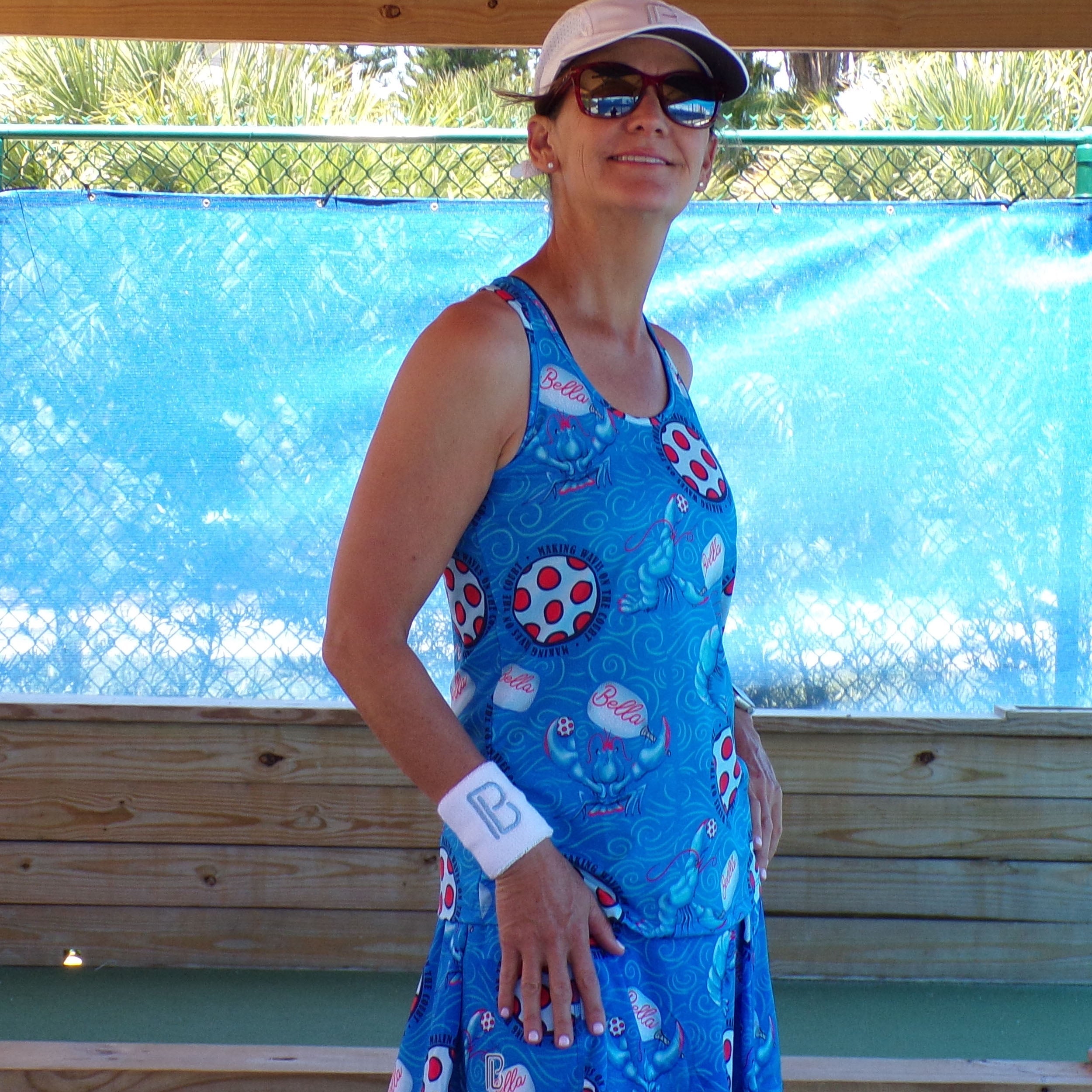 Woman wearing a blue dress with red and white patterns, sunglasses, and a cap, standing in front of a blue tarp.