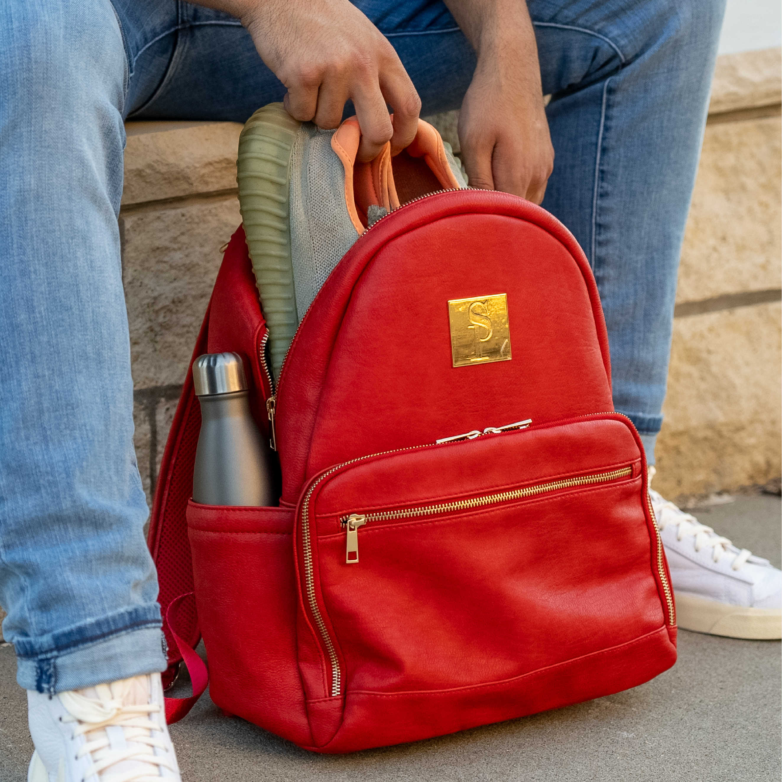 Red backpack with gold accents being held open by a person, showing a water bottle and shoes inside.