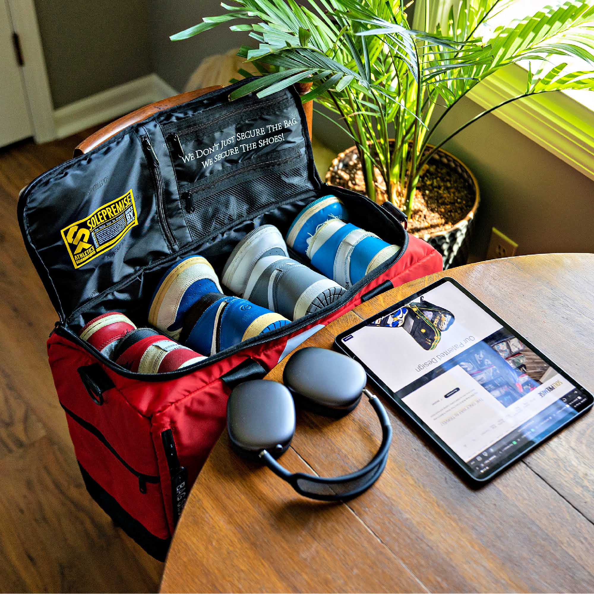 Red suitcase with shoes and a tablet on a wooden table, surrounded by plants.