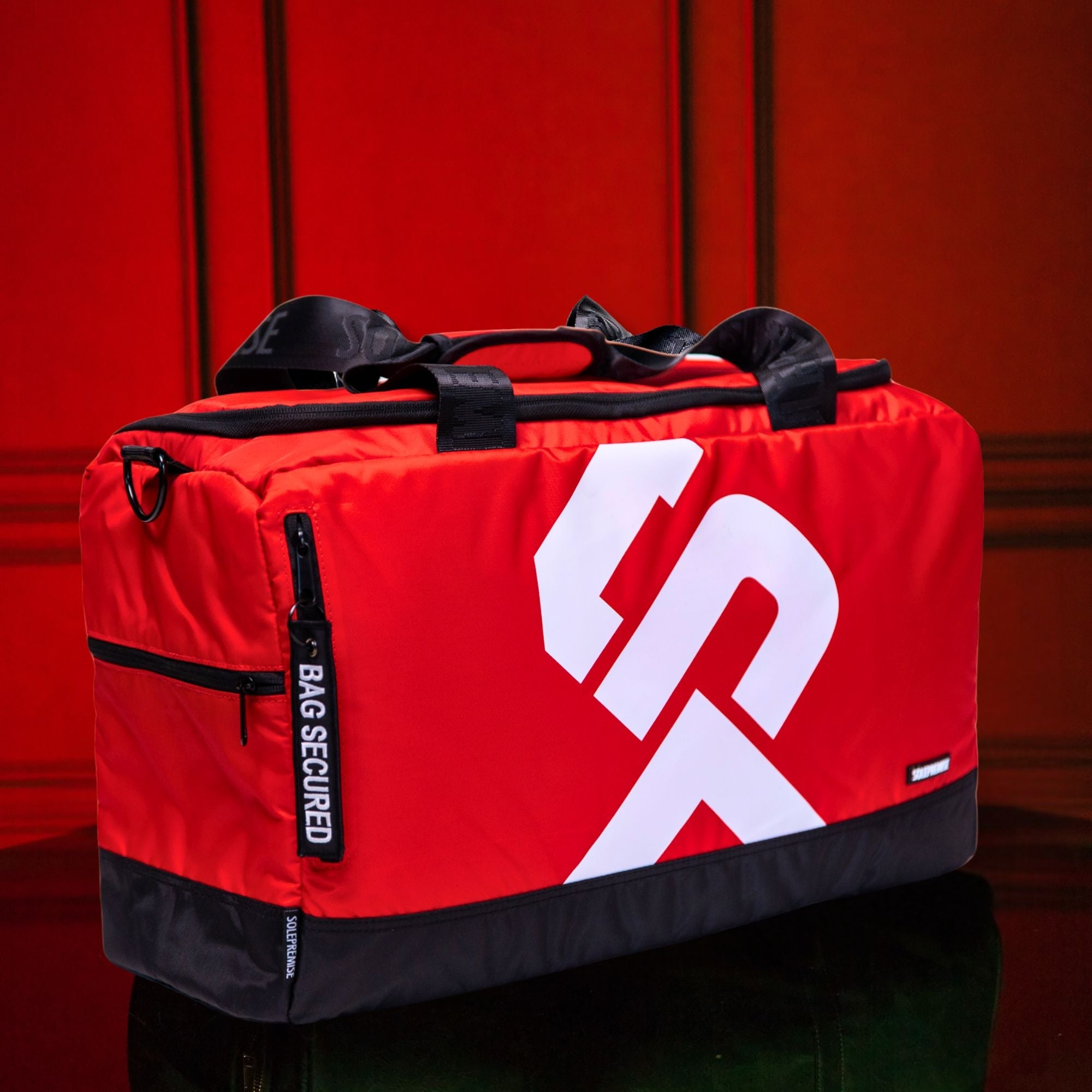 Red duffel bag with a white logo on a reflective surface with a red background