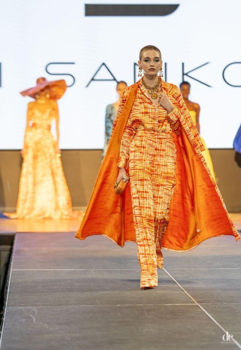 Model walking on a catwalk wearing an orange and yellow patterned outfit with a long orange drape.