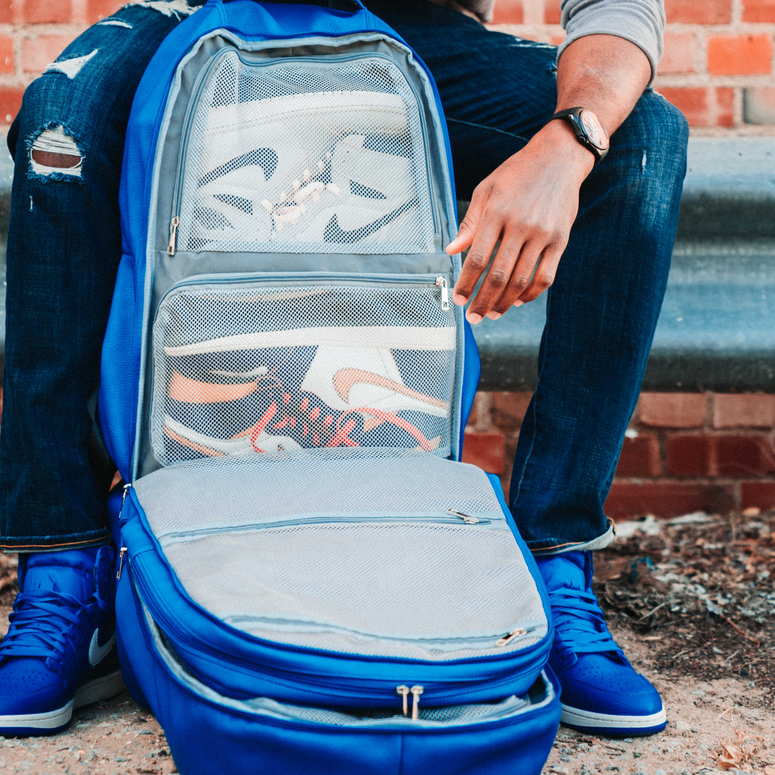 Person sitting on steps with a blue open suitcase showing white sneakers inside, against a brick wall.