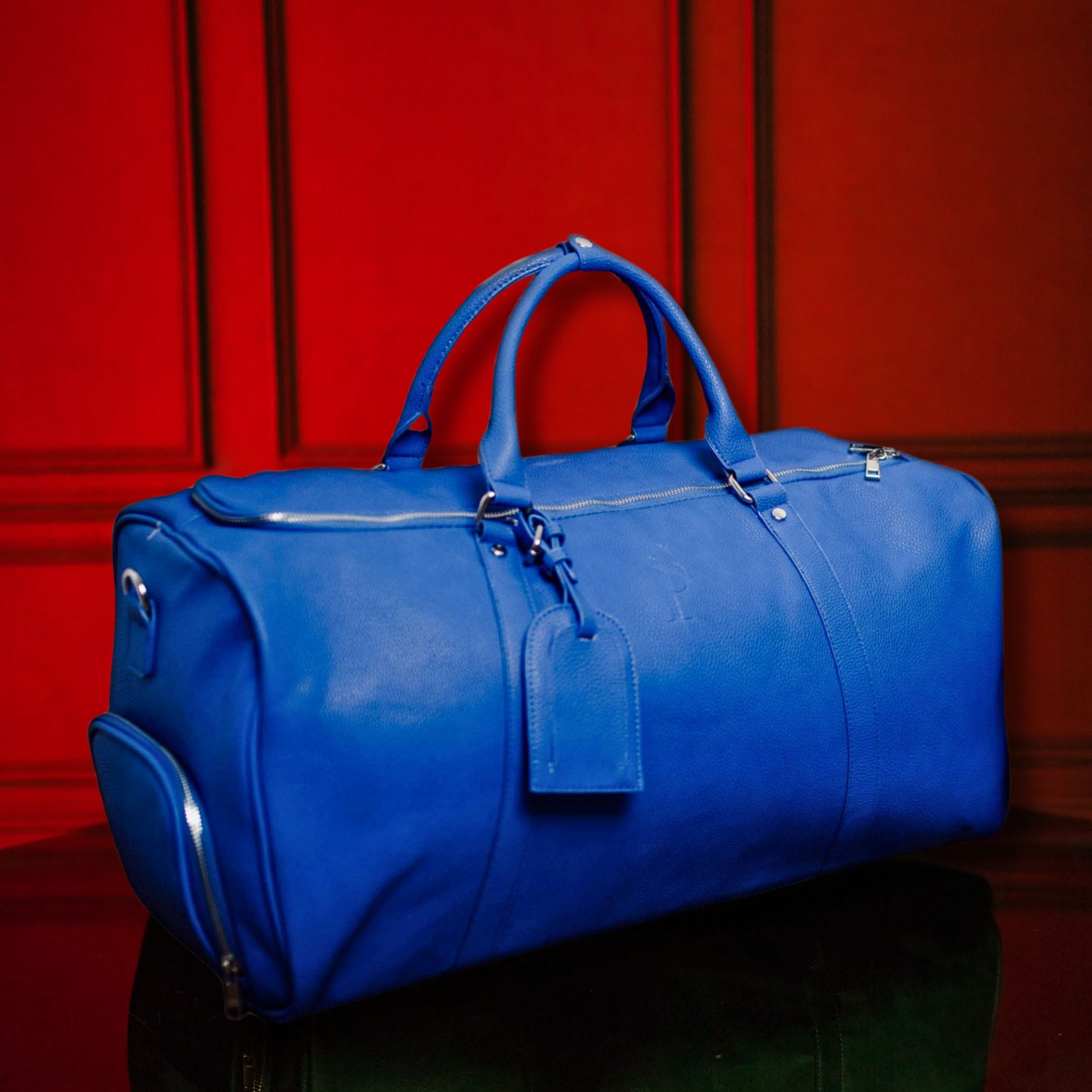 Blue duffel bag with a tag on a reflective surface against a red paneled wall.