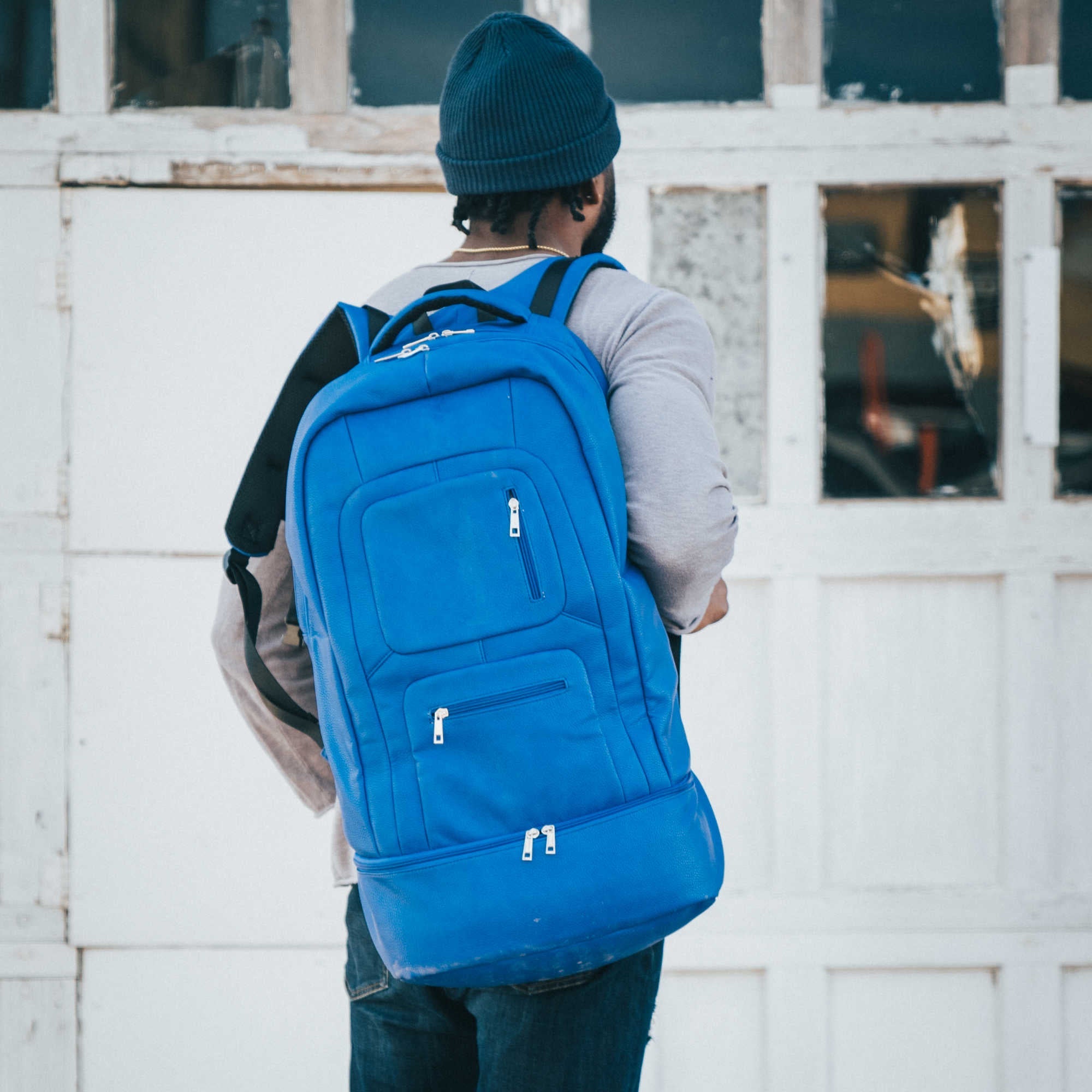 Person wearing a blue backpack in front of a white wall with windows.