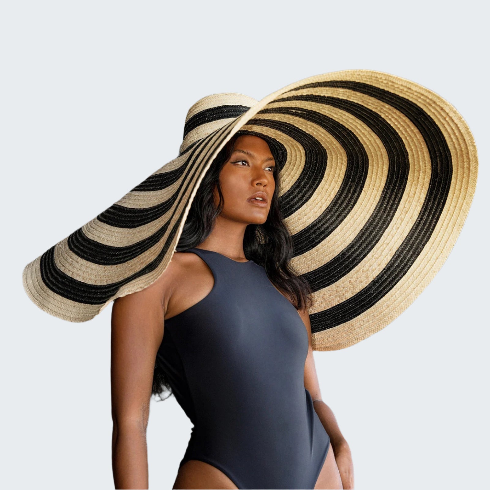 Woman wearing a black swimsuit and a large straw hat with a black and beige pattern against a rocky background.
