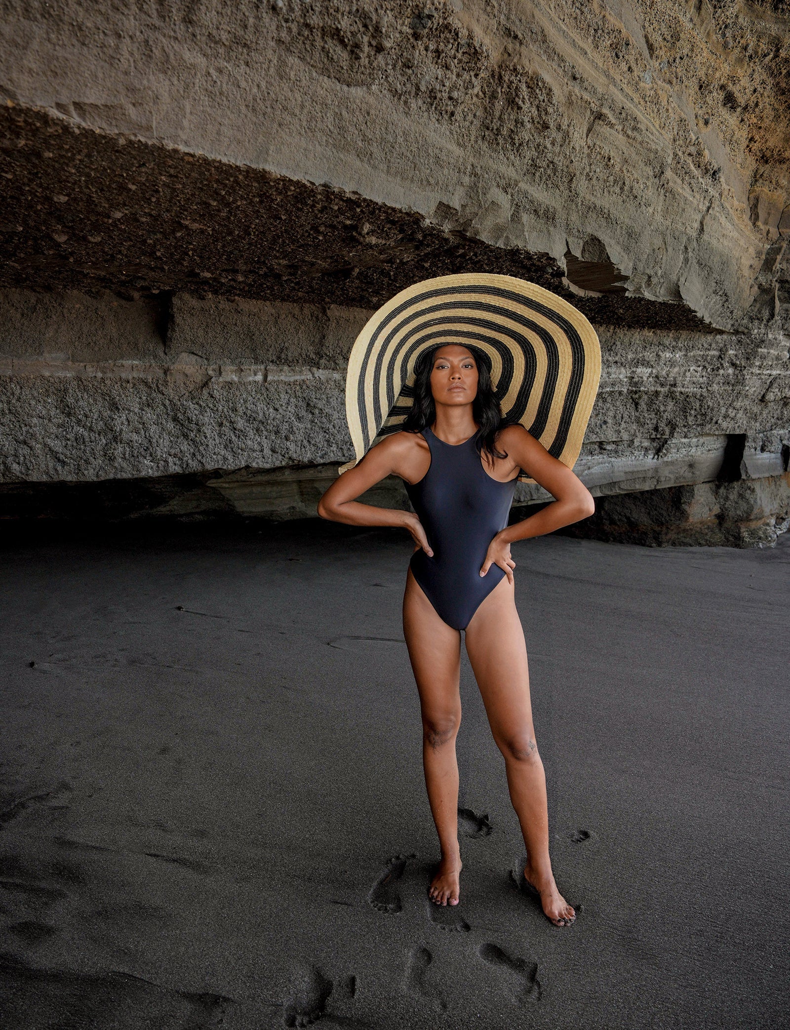 Woman in a blue swimsuit and large sun hat standing on a beach.