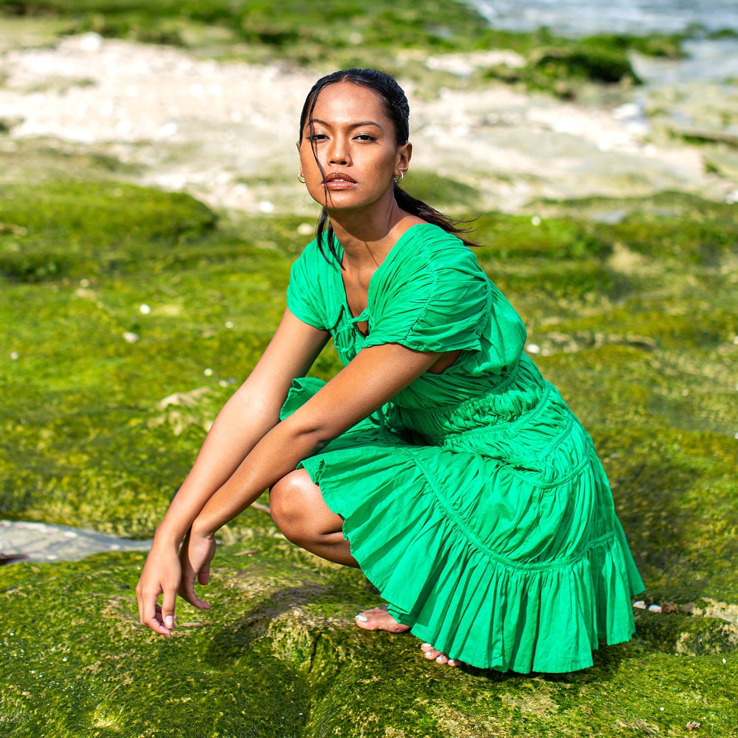 Woman in a green dress sitting on a rocky, algae-covered shore.