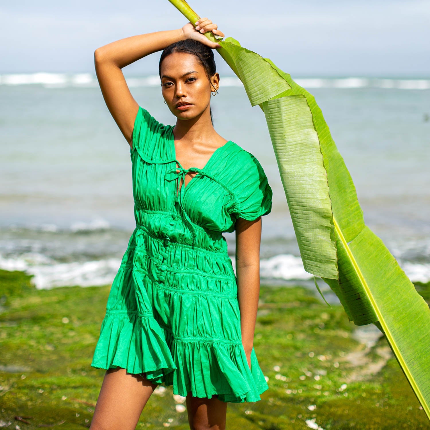 Woman in a green dress holding a large green leaf against a beach background