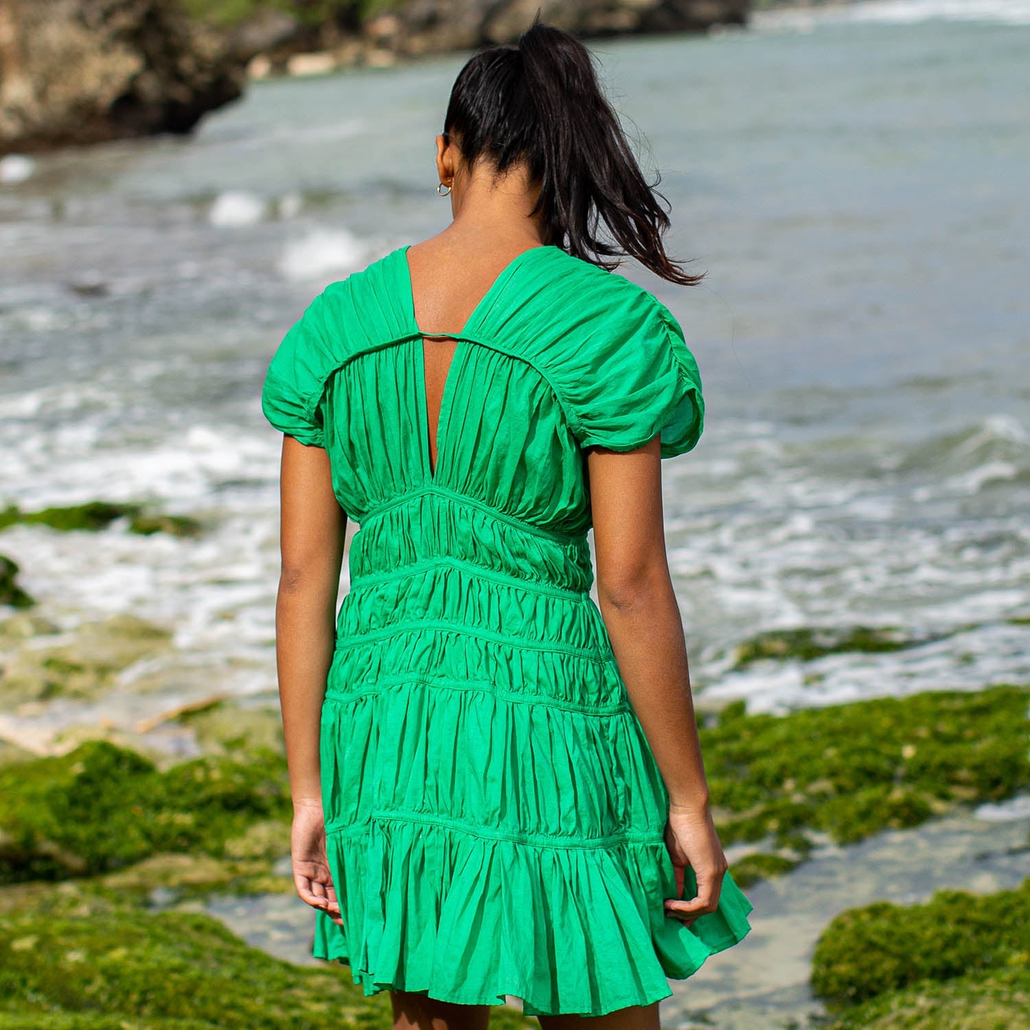 Woman in a green dress standing by a body of water with rocks and greenery.