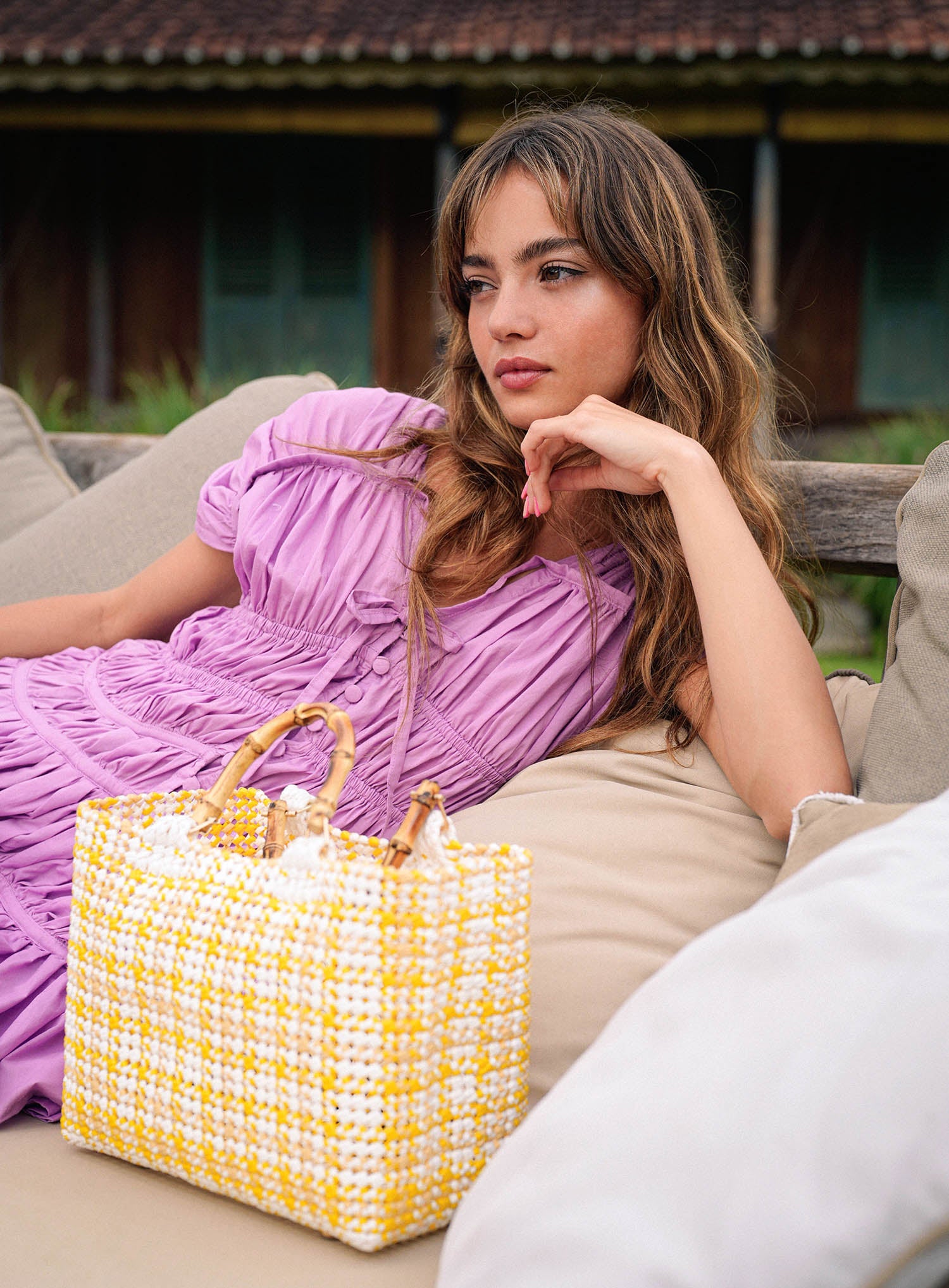 Woman in a purple dress sitting on a couch with a yellow woven bag next to her.