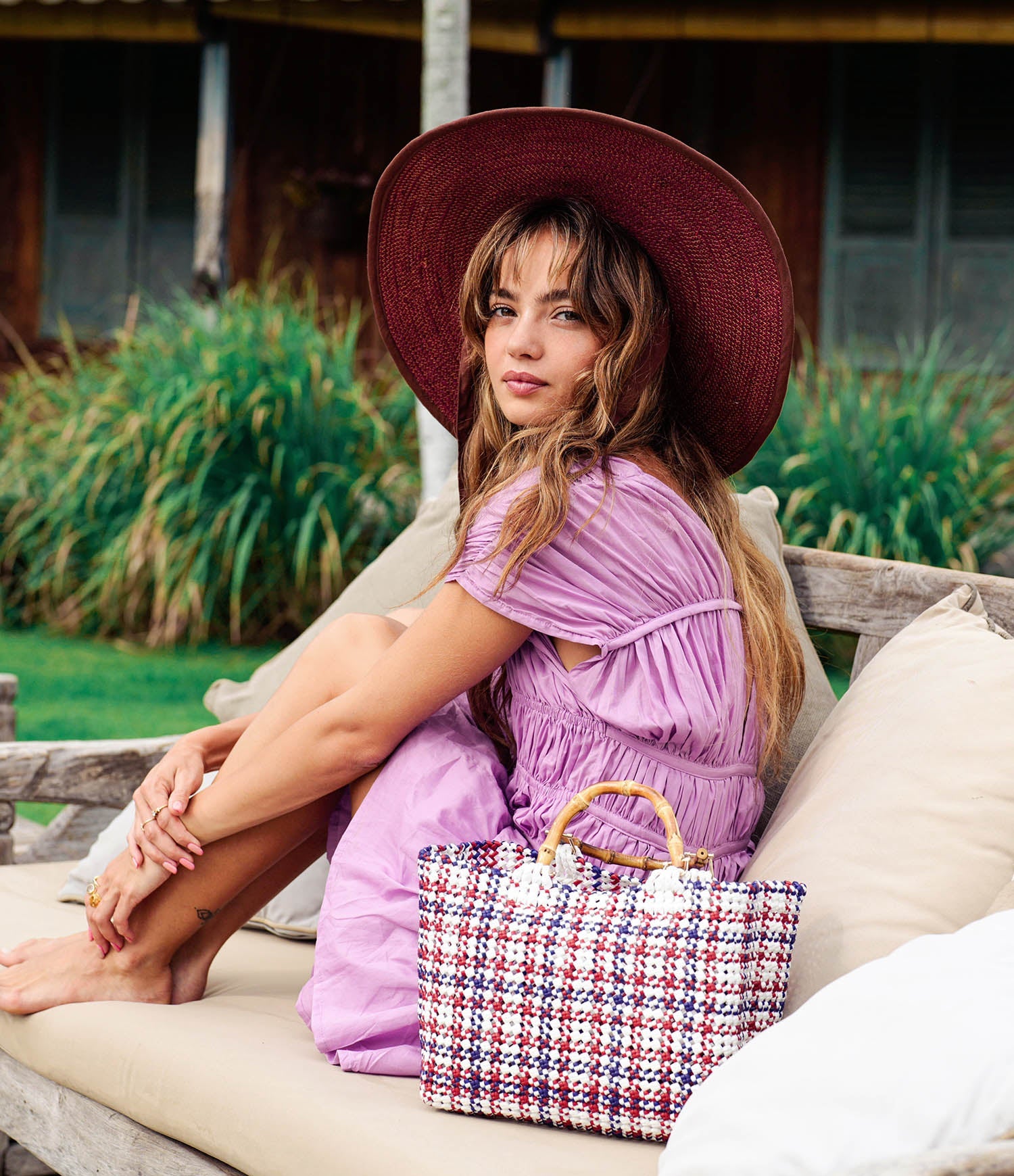 Woman in a purple dress and red hat sitting on a wooden bench with a colorful bag.