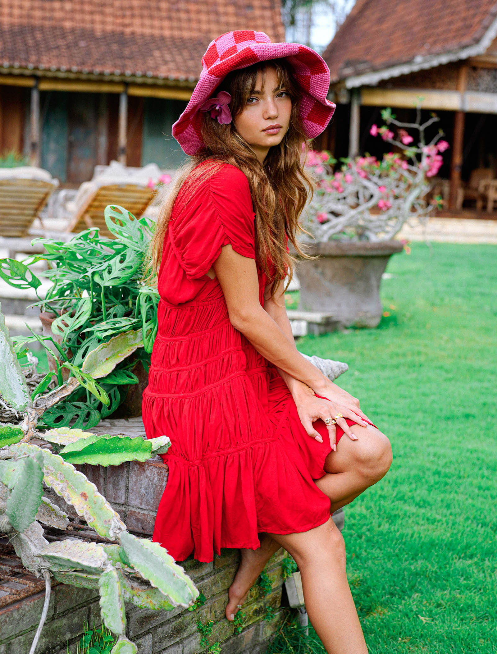 Woman in a red dress and pink hat sitting on a stone wall in a garden.