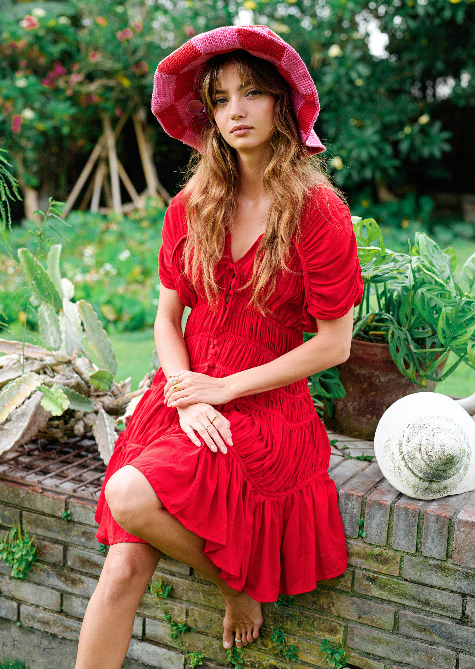Woman in a red dress and hat sitting outdoors with greenery in the background
