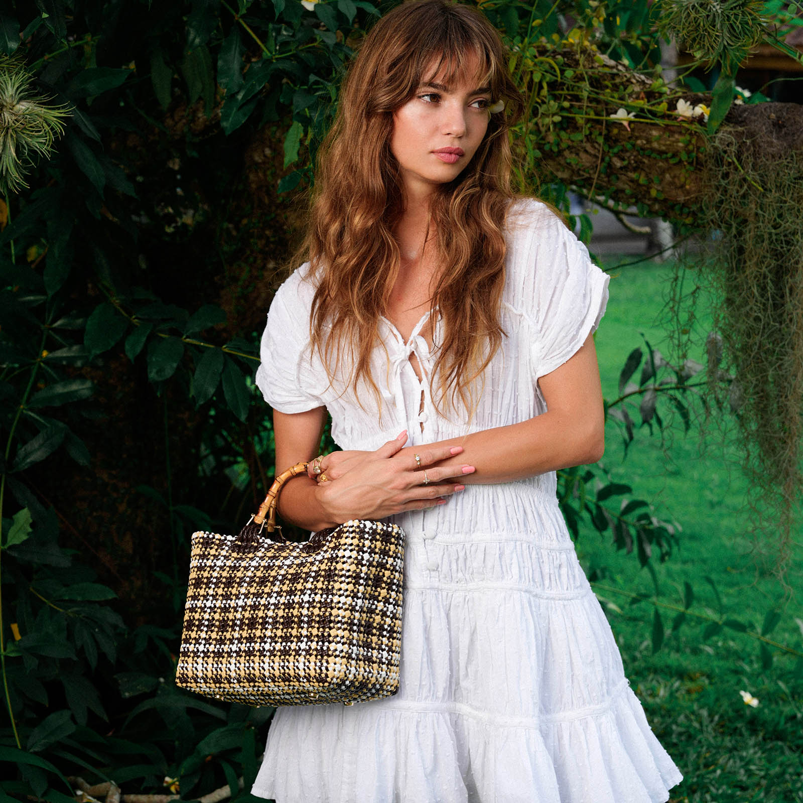 Woman in a white dress holding a woven handbag in a garden setting