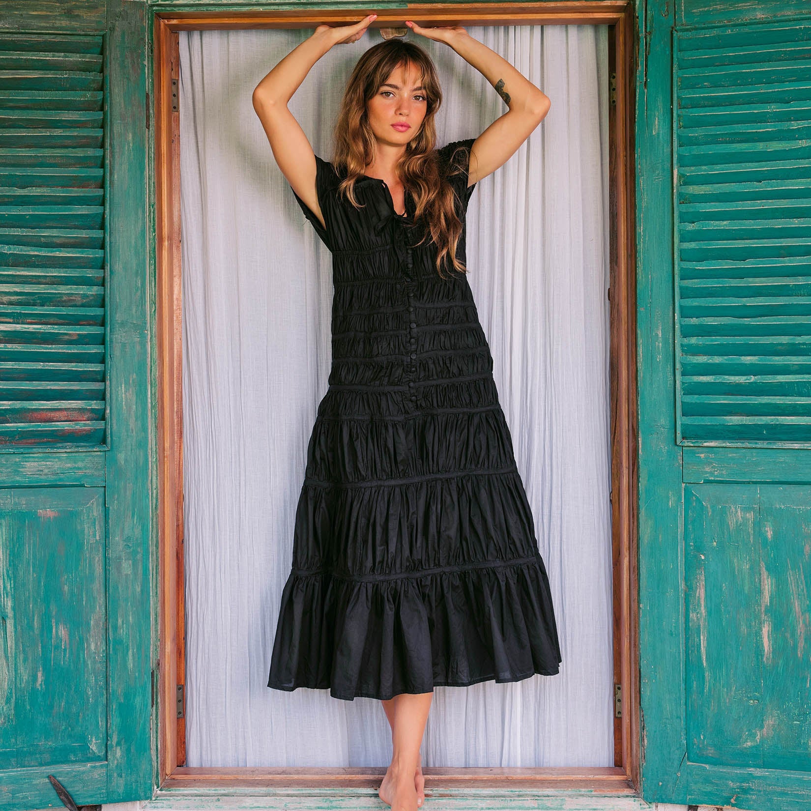Woman in a black dress standing in a doorway with green shutters.