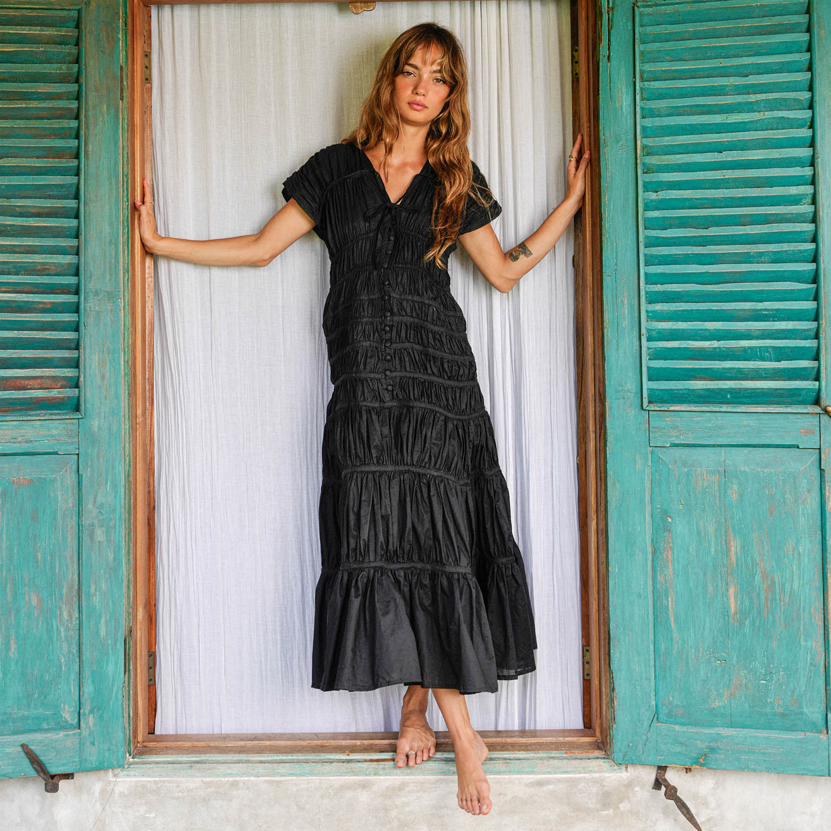 Woman in a black dress standing in front of a rustic door with teal shutters.