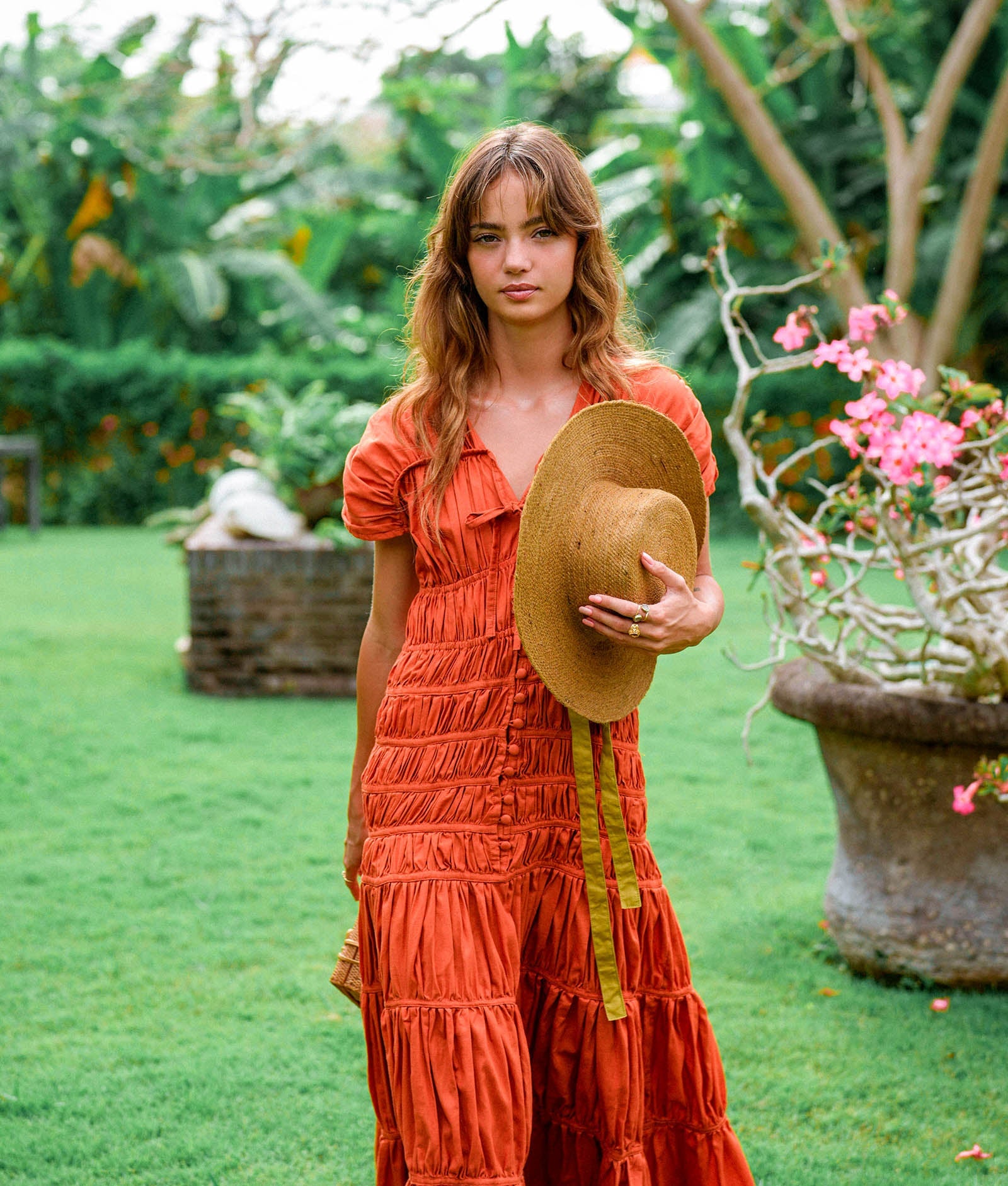 Woman in an orange dress holding a straw hat in a garden setting