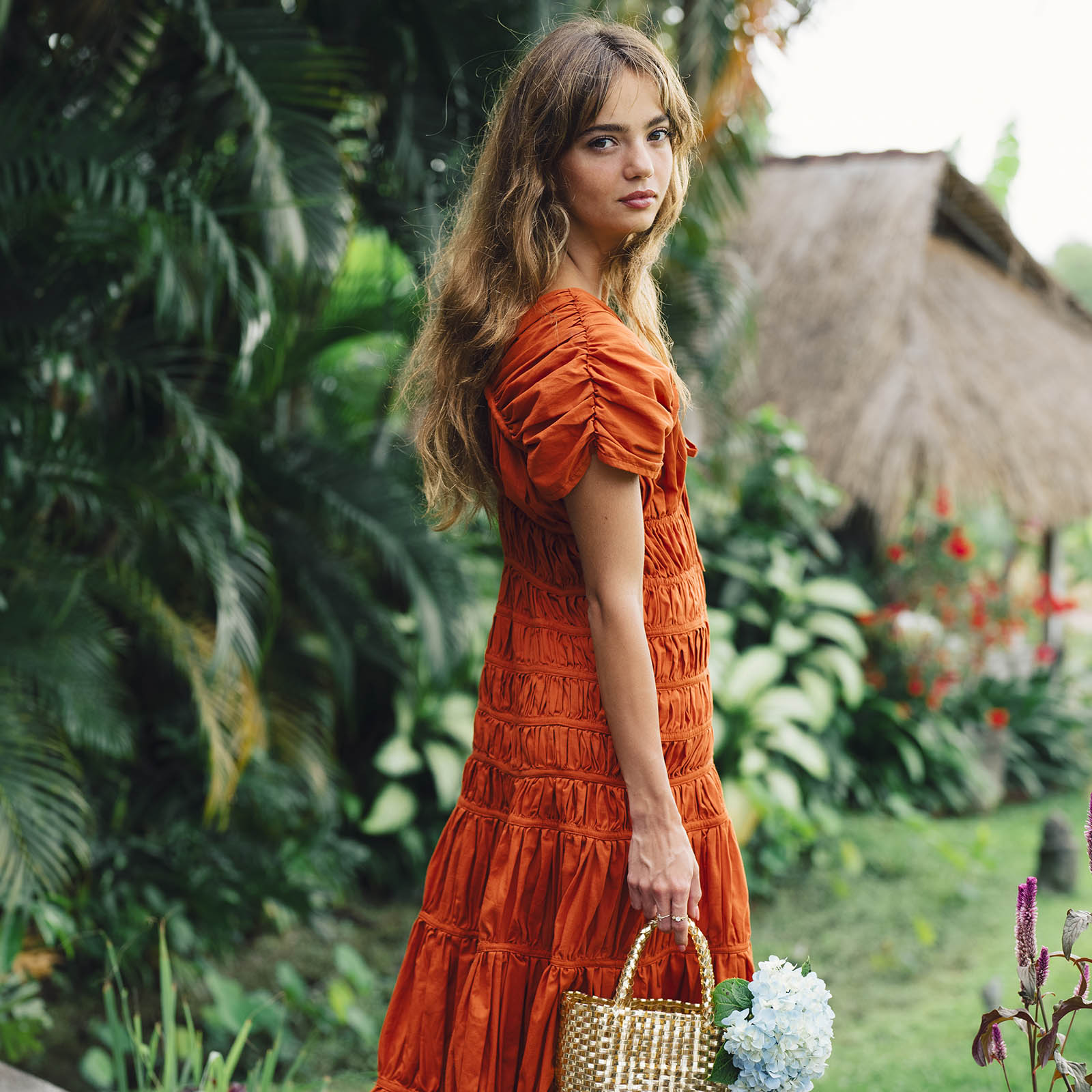Woman in an orange dress standing in a garden with greenery and flowers.