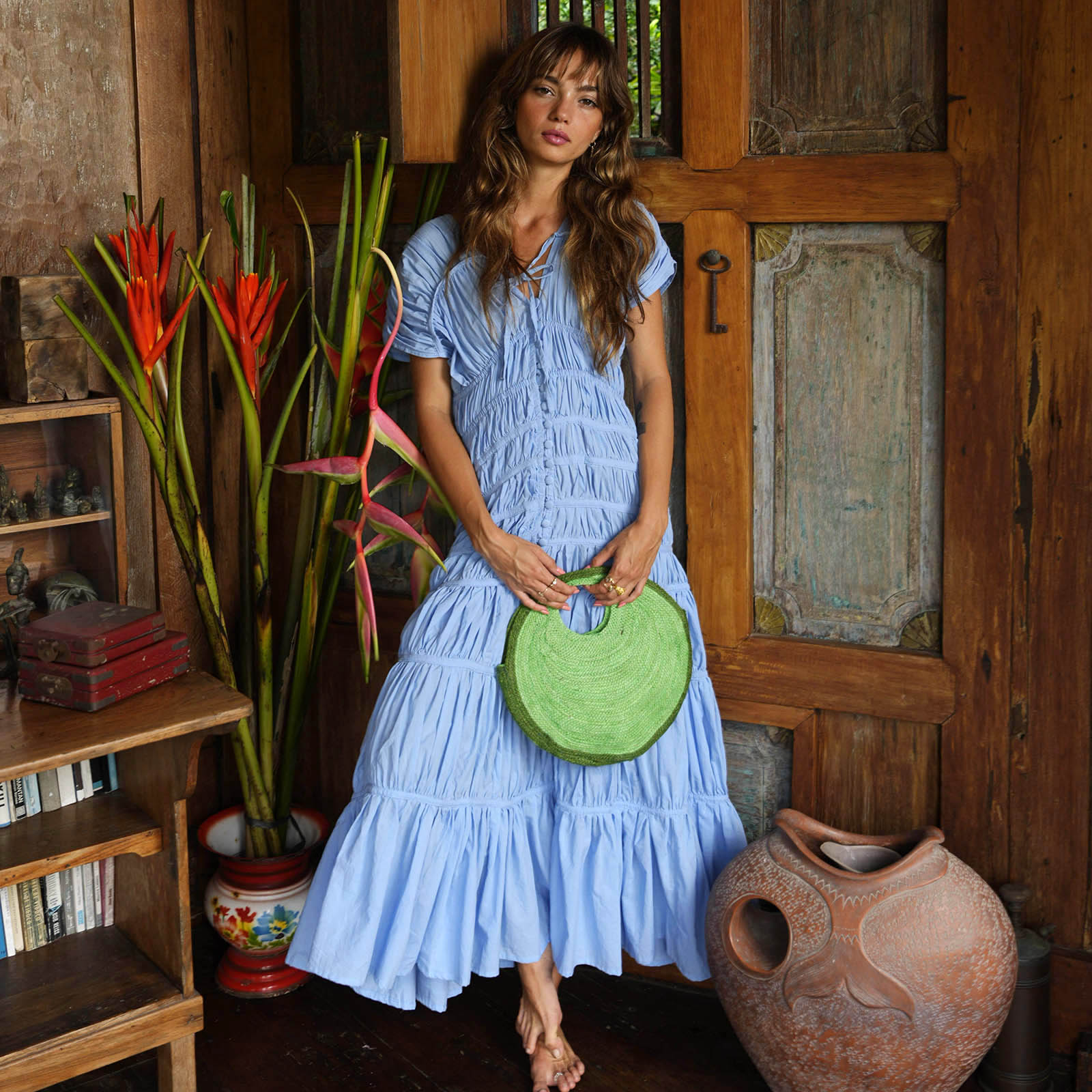 Woman in a blue dress holding a green bag in a rustic indoor setting with wooden furniture and plants.